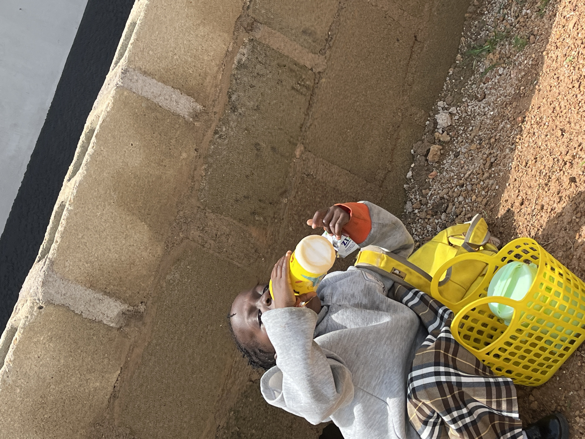In Barakin Akawo, Nigeria, a young child is seated outdoors, leaning against a textured concrete wall. The child, wearing a grey hooded sweatshirt and plaid trousers, is drinking from a yellow bottle. A bright yellow plastic basket containing a light green container and other items sits next to the child. The ground is covered in dirt and small stones. The scene is illuminated by natural daylight.