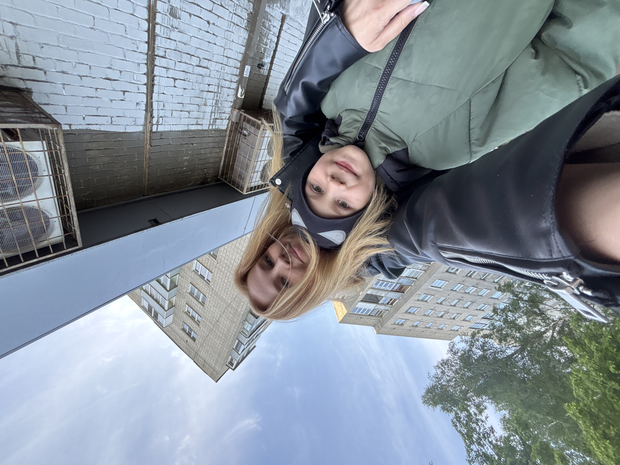 A selfie-style, rotated image shows two females, one adult and one child, against an urban background of buildings and sky. The adult female has blonde hair and is wearing a black jacket. The child is wearing a black and white patterned headband, and a green jacket. The image is taken from a low angle.
The background includes apartment buildings, with visible windows and balconies, and a partly cloudy sky. Trees are visible in the lower right portion of the image. The location appears to be Rusanovo, Russia, indicated by the architectural style and urban setting typical of the region. Additional visual clues, such as air conditioning units and brickwork, suggest the photo was taken near a residential area.