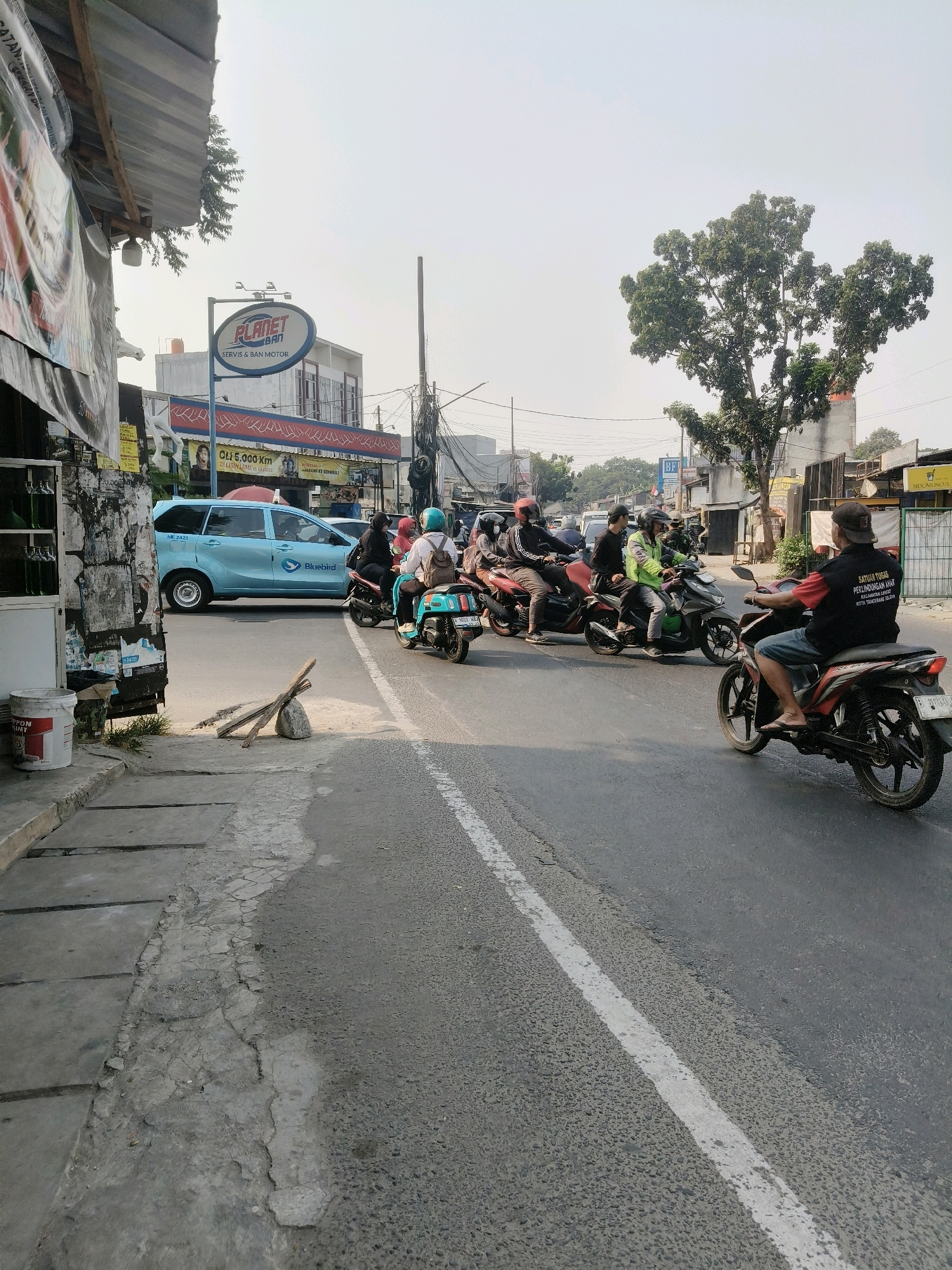 The image depicts a street scene in South Tangerang, Indonesia, on a cloudy day. A light blue Bluebird taxi is stopped in the left lane of a two-lane road, with several motorcycles in the right lane, some of which are in motion. One motorcycle in the foreground on the right has a rider wearing a black shirt with white text and a dark baseball cap, riding a red and black motorcycle. In the distance, there are buildings with signage, including a prominent "PLANET BAN" sign for motorcycle tires and service. Trees and utility poles are visible along the street. The road surface is asphalt, with a white line marking the lanes. A concrete sidewalk runs along the left side of the road, showing signs of wear and tear. Various posters and advertisements are affixed to a structure on the left.