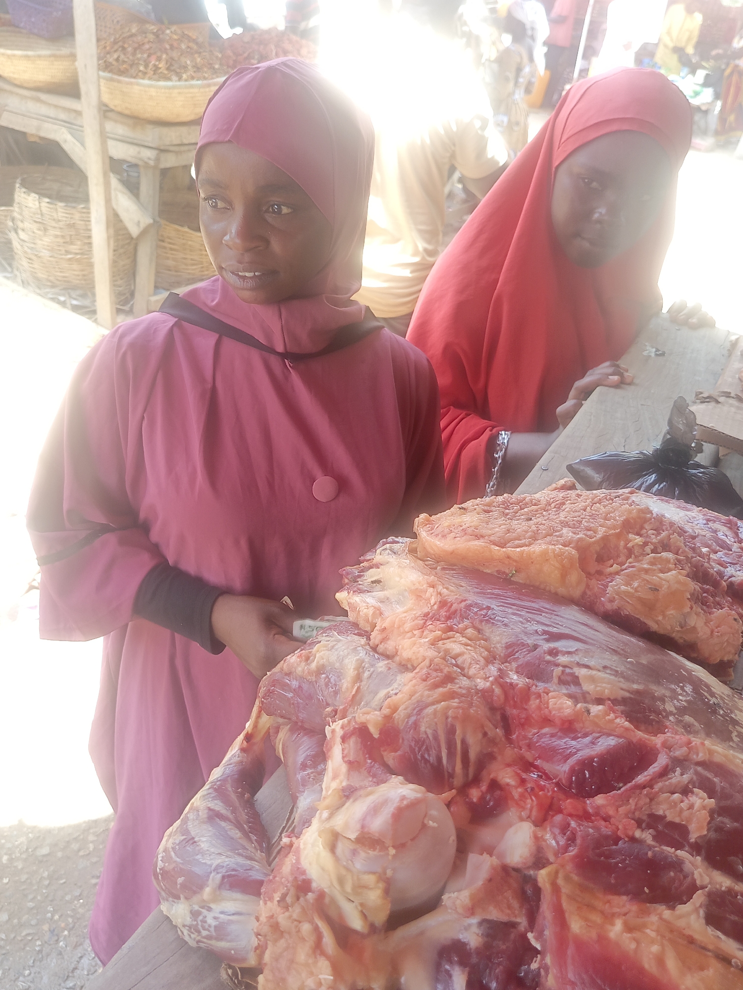 The image shows two women at an outdoor meat stall in Akwanga, Nigeria. The woman in the foreground is wearing a pink head covering and a matching long-sleeved dress with black cuffs. She is holding currency in her hands and looking to the left. Behind her, another woman is wearing a red head covering. In front of them is a large display of raw meat on a wooden surface. A black plastic bag rests on top of the meat. In the background, there is a market scene with baskets and other goods visible. The lighting appears bright and sunny.