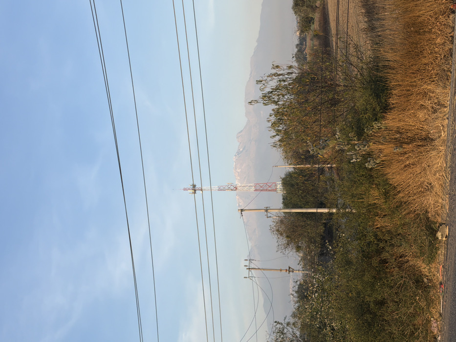 The image depicts a static, outdoor landscape scene in the city of Santa Ana Xalmimilulco, Mexico, observed under a clear, light blue sky with minimal atmospheric haze. The foreground features a dark grey paved surface at the very bottom, transitioning into a strip of dry, golden-brown grasses and scattered green shrubs that span the width of the frame. A small piece of light-colored debris is visible on the paved surface near the bottom left.
Horizontally traversing the mid-ground are five or more dark power lines, partially obscuring the view. Below these lines, several wooden utility poles are visible, primarily on the left. A tall, red and white lattice communication tower stands prominently in the center-left, equipped with multiple antennae arrays near its apex. To the right and behind the utility poles and tower, the landscape consists of varied vegetation, including dense green and brown trees and shrubs, and areas of dry, light-brown agricultural land. Faint outlines of distant urban or semi-urban structures are discernible through the atmospheric haze below the mountain range.
The background is dominated by a distant, large mountain range with visible snow or ice patches on its peaks, particularly on the central and left portions of the range. The mountains appear somewhat muted due to atmospheric haze. No individuals or animals are present. The scene is illuminated by natural light, suggesting either early morning or late afternoon, with a general absence of harsh shadows.