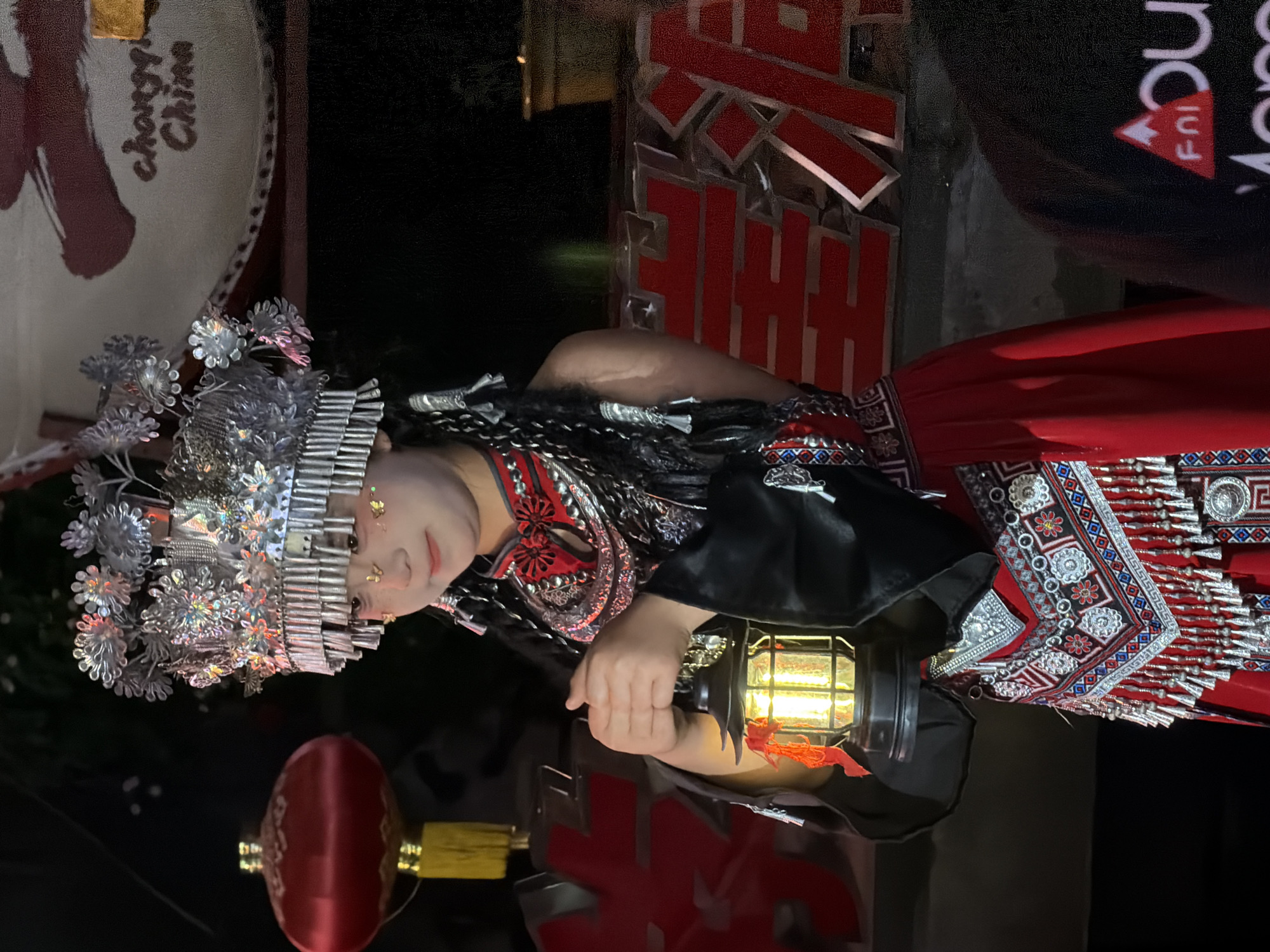 A young girl, likely of Han Chinese ethnicity, stands outdoors at night, holding a lit lantern. She wears a traditional, ornate costume featuring a headdress adorned with silver embellishments and red accents, and a red dress with intricate embroidered patterns and silver fringe. Her hair is styled in braids with silver ornaments. She has glitter or face paint on her cheeks, resembling small butterflies. In the background, a large red lantern hangs to the left, and a sign that reads "chongqi China" is visible to the right, suggesting the location is in Chongqing, China. Red Chinese characters are also visible in the background. The overall scene appears festive, possibly part of a cultural event or performance.