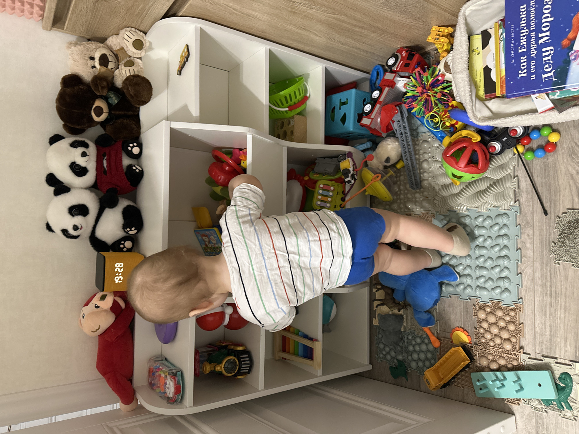 A baby is captured in a child's play area, leaning into a white cubby shelf filled with toys. The baby, dressed in a striped long-sleeve shirt, blue shorts, and white socks, appears to be exploring or reaching for items within the shelf. The floor is covered with various sensory play mats, featuring different textures like bumps and ridges, designed for tactile exploration.
The room is rich with children's toys and accessories. To the left, a collection of plush animals includes several brown bears, three black and white panda bears, and a red monkey. The white storage unit contains a green basket, a blue shape sorter, a colorful xylophone, and various small plastic toys. Scattered on the floor next to the shelf are more toys, such as a red toy fire truck, a yellow dump truck, a bright green and red rattle, a multi-colored spiky ball, and other small playthings. A blue plush animal also lies on the floor.
In the upper right corner, a white fabric basket holds several children's books. Visible text on one of the book covers in Cyrillic reads "М. КРИСТИНА БАТЛЕР" (M. Kristina Butler) and a title that translates to "How Hedgehog and his friends helped Ded Moroz (Grandfather Frost)." Another fragment of text visible says "ведут ко сну" (lead to sleep). A digital clock on a shelf to the left displays "19:28," indicating the time is 7:28 PM. The overall scene suggests an evening playtime session in a well-equipped child's room or dedicated play space.