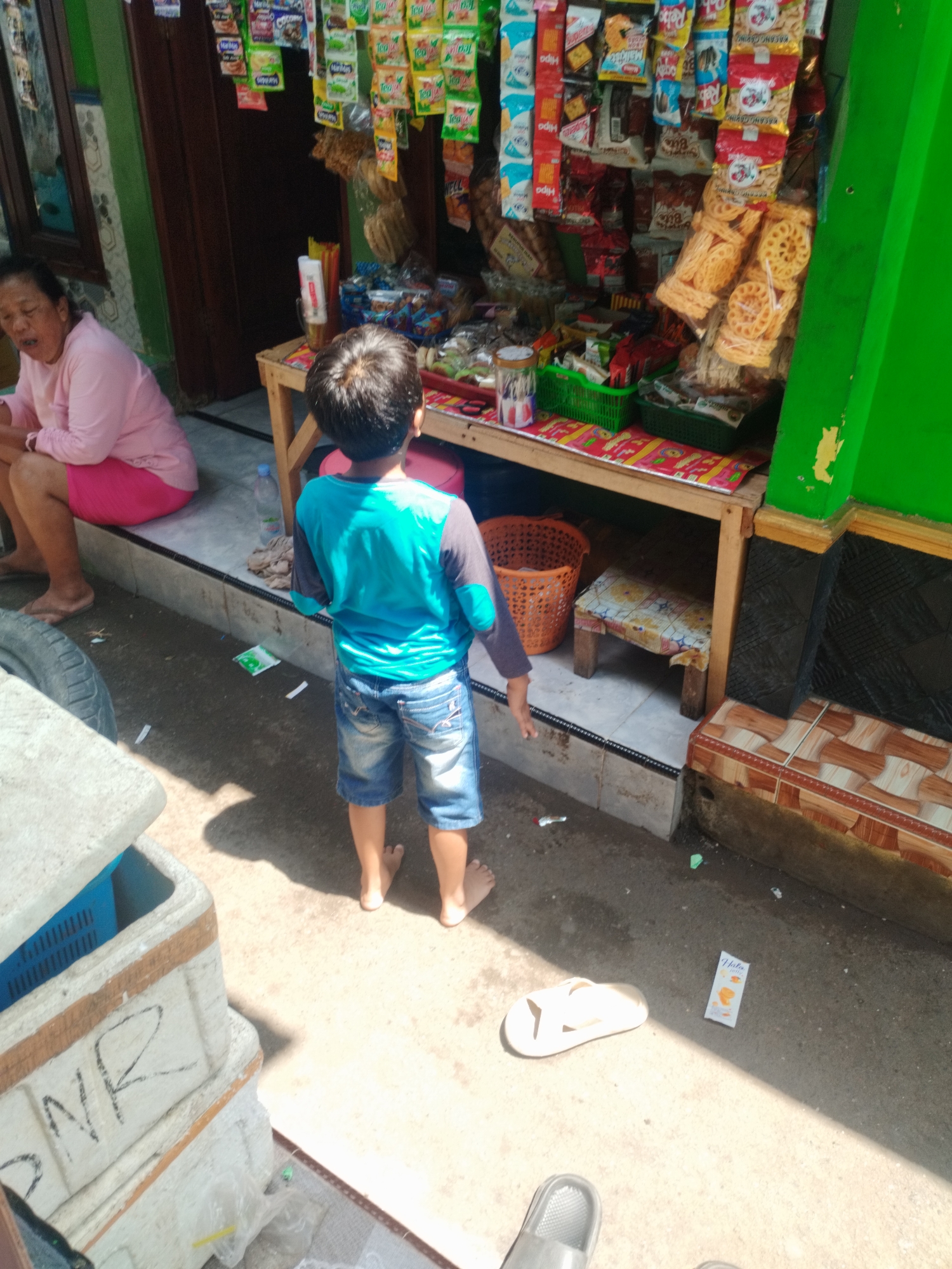 A medium shot captures a street-level view of a small outdoor market stall in Kelurahan Cilacap, Kecamatan Cilacap, Indonesia. Sunlight casts distinct shadows across the scene. In the foreground, a young boy, facing away from the camera, stands barefoot in a bright blue t-shirt and denim shorts. To the left, an adult woman in a pink shirt and pink shorts sits with her legs extended. The market stall is filled with an assortment of packaged snacks and goods displayed on shelves. A low wooden table holds a woven basket and a small stool covered in a patterned cloth. Various items, including plastic bags and a water bottle, are scattered on the ground. In the far right of the frame, a bright green wall of the structure is visible.