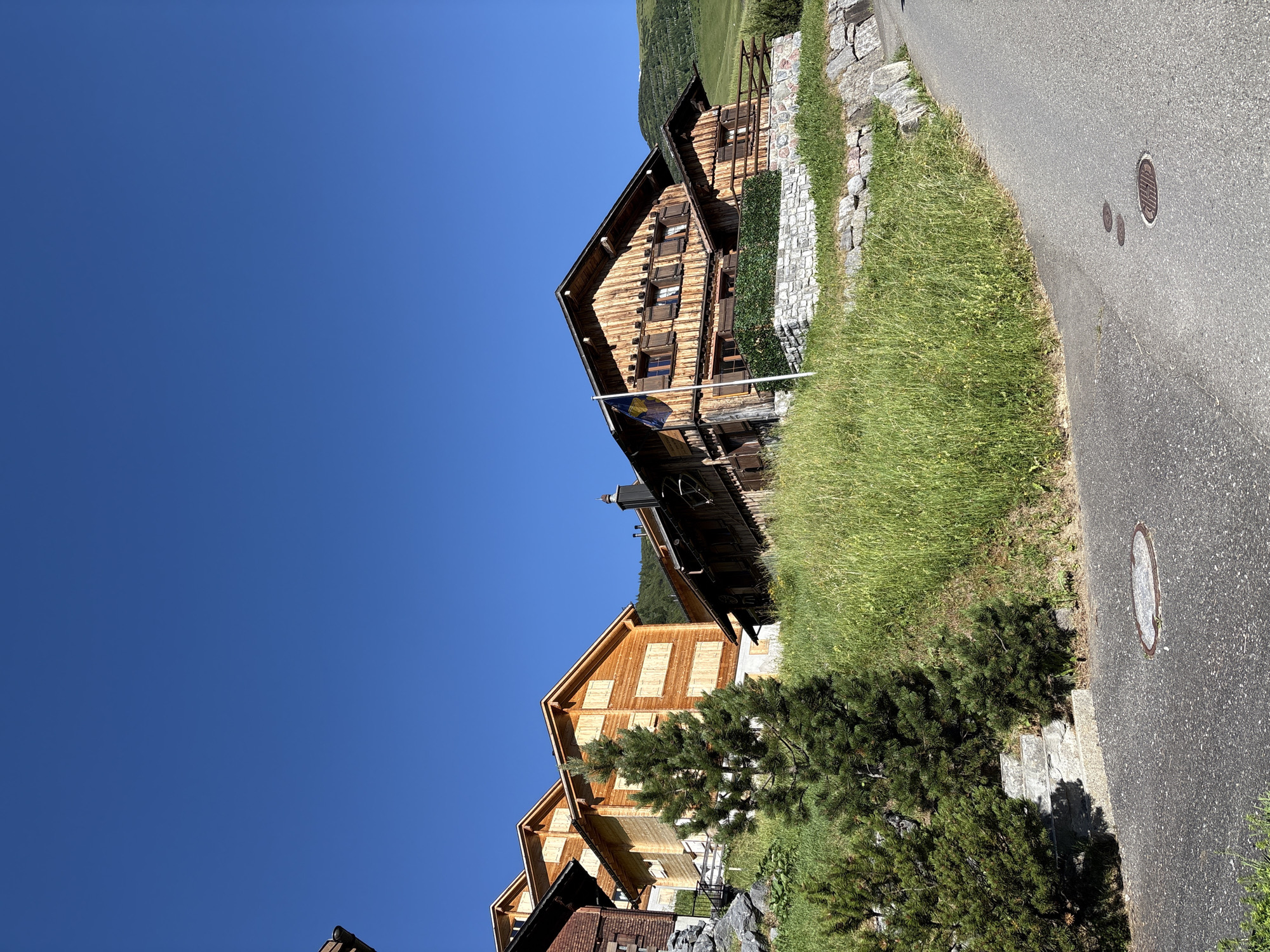 The image captures a sun-drenched, upward view of a collection of traditional wooden chalets nestled on a grassy slope in Malbun, Liechtenstein. The sky is a clear, vibrant blue. Several multi-story chalets with wooden facades and shutters are prominent. One chalet features a flag with blue and yellow stripes. The buildings are set against a backdrop of rolling green hills. In the foreground, a paved road curves upwards, with a manhole cover visible on its surface. Lush green grass and several evergreen trees populate the slope between the road and the chalets. The scene depicts a peaceful, mountainous village setting.