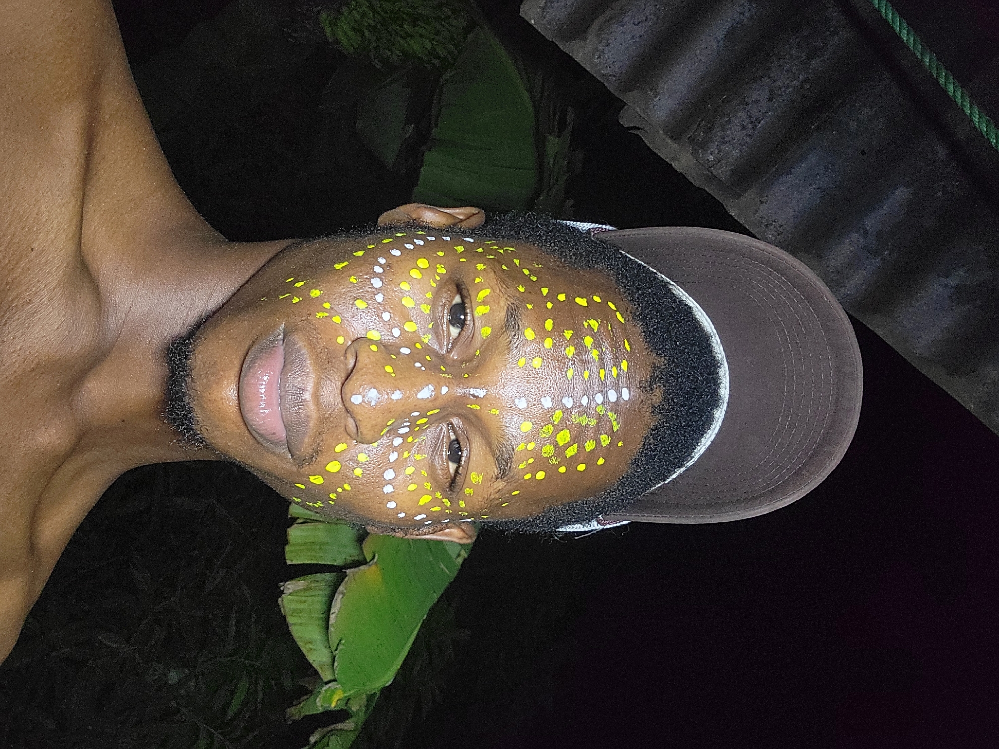A close-up of a person's face with yellow and white dots painted on it. The person has dark skin, a beard, and is wearing a baseball cap. A banana leaf is visible in the background. The location is Nkpor, Nigeria, based on contextual knowledge. The picture appears to be taken at night or in low light.