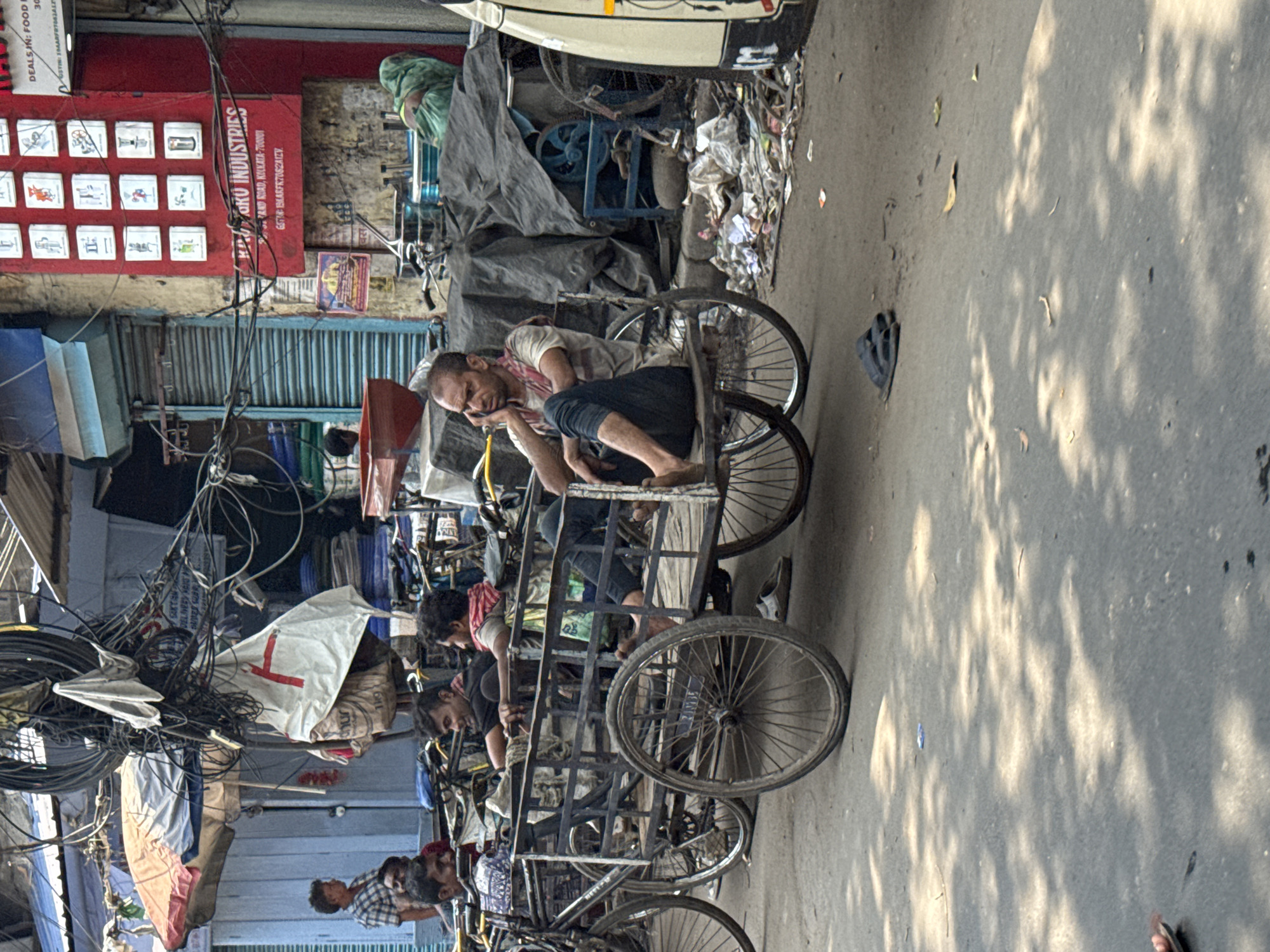 The image depicts a street scene in Kolkata, India, featuring several rickshaws and people. A man is reclining within a rickshaw, appearing to be resting. Other individuals are seen around the rickshaws, with varying levels of engagement. The street is cluttered with wires overhead and various objects and materials scattered around. The scene is illuminated by sunlight, casting shadows on the pavement. A red and white sign is visible in the background, indicating "OKO INDUSTRIES" on Strand Road, Kolkata-700001. The sign also includes a GSTIN number, suggesting a commercial establishment. The overall impression is that of a busy, somewhat rundown street scene characteristic of urban India.