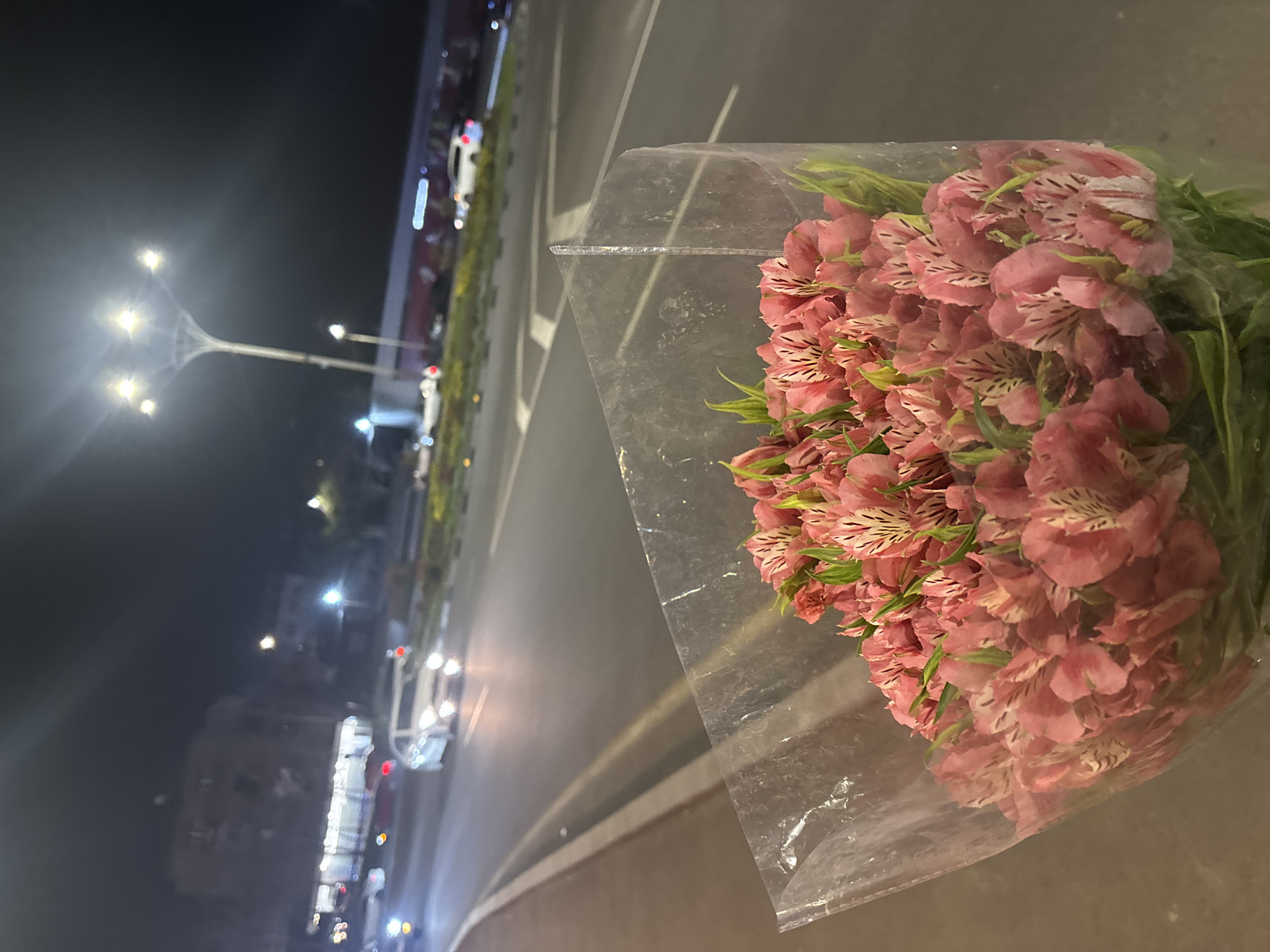 A bouquet of pink alstroemeria flowers wrapped in clear plastic sits on a surface at night in Yerevan, Armenia. A street stretches out from the right foreground into the background, where streetlights illuminate the road and buildings. Cars are visible on the street in the distance.
