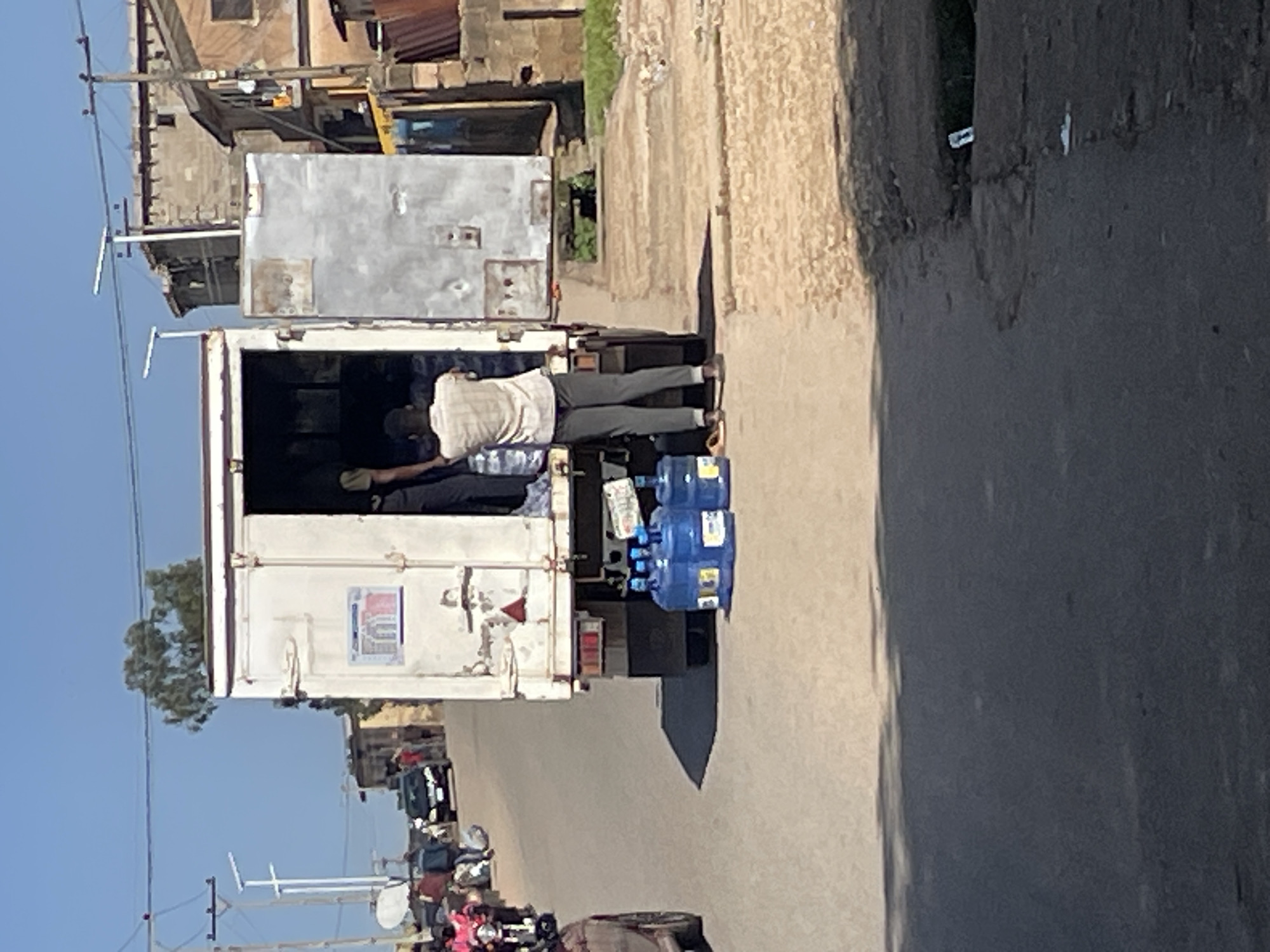 The image shows a street scene in Jos, Nigeria, with a large white truck in the foreground. The truck's rear doors are open, and two men are visible inside and near the back of the truck. One man, wearing a light-colored shirt and dark pants, is standing by the open doors of the truck, appearing to be unloading or loading something. Another person, wearing dark clothing, is partially visible inside the truck. Several large blue water cooler jugs are stacked on the ground behind the truck. In the background, there are buildings, a black car, and several motorcycles, suggesting a typical urban environment. The sky is clear and blue, and the overall lighting indicates it is daytime.