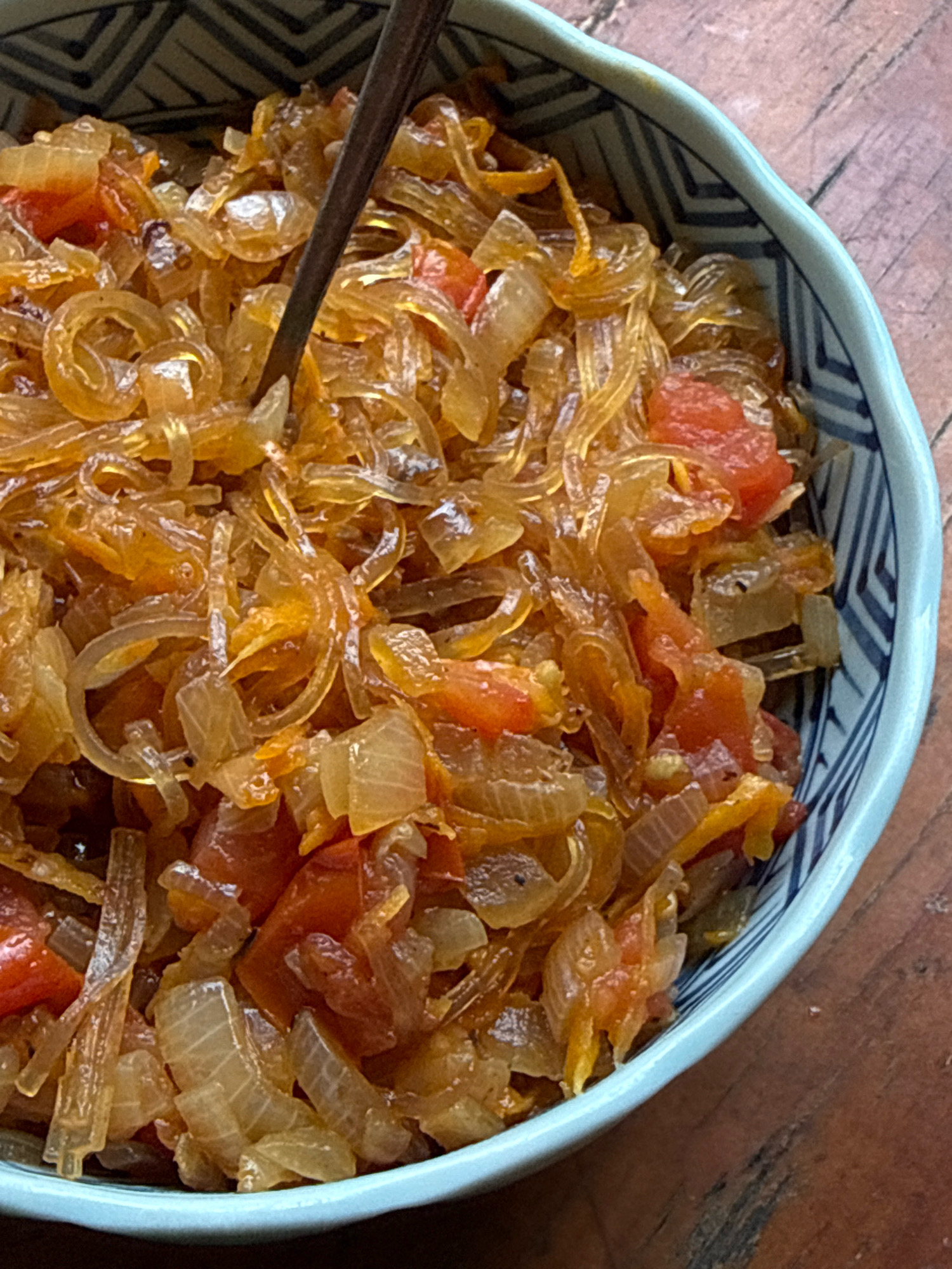 A close-up, overhead shot shows a bowl filled with cooked noodles, tomatoes, and onions, served on a wooden table. A dark utensil is partially submerged in the food. The bowl is light blue with a dark blue patterned rim. The food appears to be a savory dish, possibly stir-fried noodles. The lighting is warm and natural, highlighting the textures of the food and the wood grain of the table. The scene appears to be indoors, with no specific details about the city of Hiệp Thạnh, Vietnam.