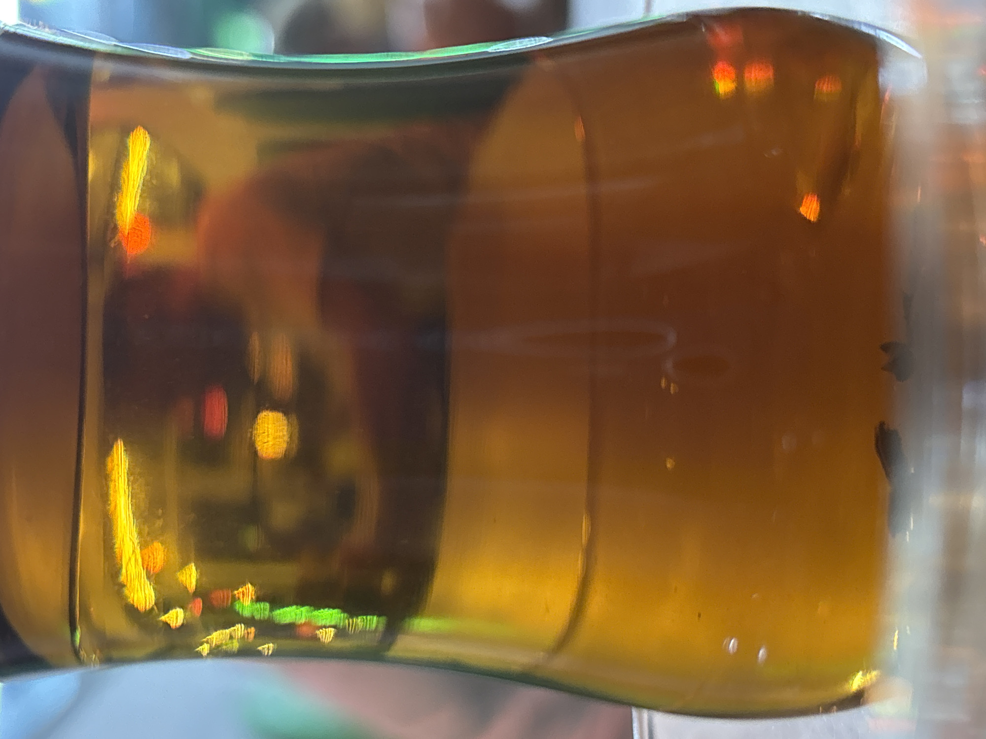 The media file presents a close-up, horizontal shot of a translucent glass container, likely a bottle or a drinking glass, filled with an amber-colored liquid. The liquid fills approximately two-thirds of the container, leaving headspace above. Vibrant, distorted reflections of various light sources are prominently featured, appearing as bright shapes in shades of yellow, orange, red, and green, visible on the glass surface and refracted through the liquid. These reflections suggest an indoor environment with artificial lighting, such as a bar or restaurant setting, in Kuta Selatan, Indonesia.
The background is out of focus, showing only blurred colors and shapes. Within the amber liquid on the right side, a few small, dark, irregular specks, possibly sediment or debris, are visible, alongside some tiny, bright, circular highlights which may be bubbles or further reflections. No people are present, and no specific activity is taking place; the image is a static shot of the container. There is no visible text. The time of day is not explicitly indicated but implied to be evening or indoors.