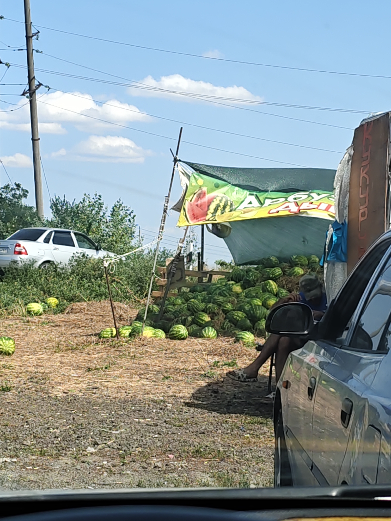 A street vendor is seen selling watermelons in Yagodinka, Russia. The vendor is sitting on a chair next to a large pile of watermelons. A banner advertising the watermelons hangs above the stall. A silver car is parked nearby, and another car is partially visible in the foreground. The scene is set outdoors on a sunny day, with trees and power lines visible in the background.