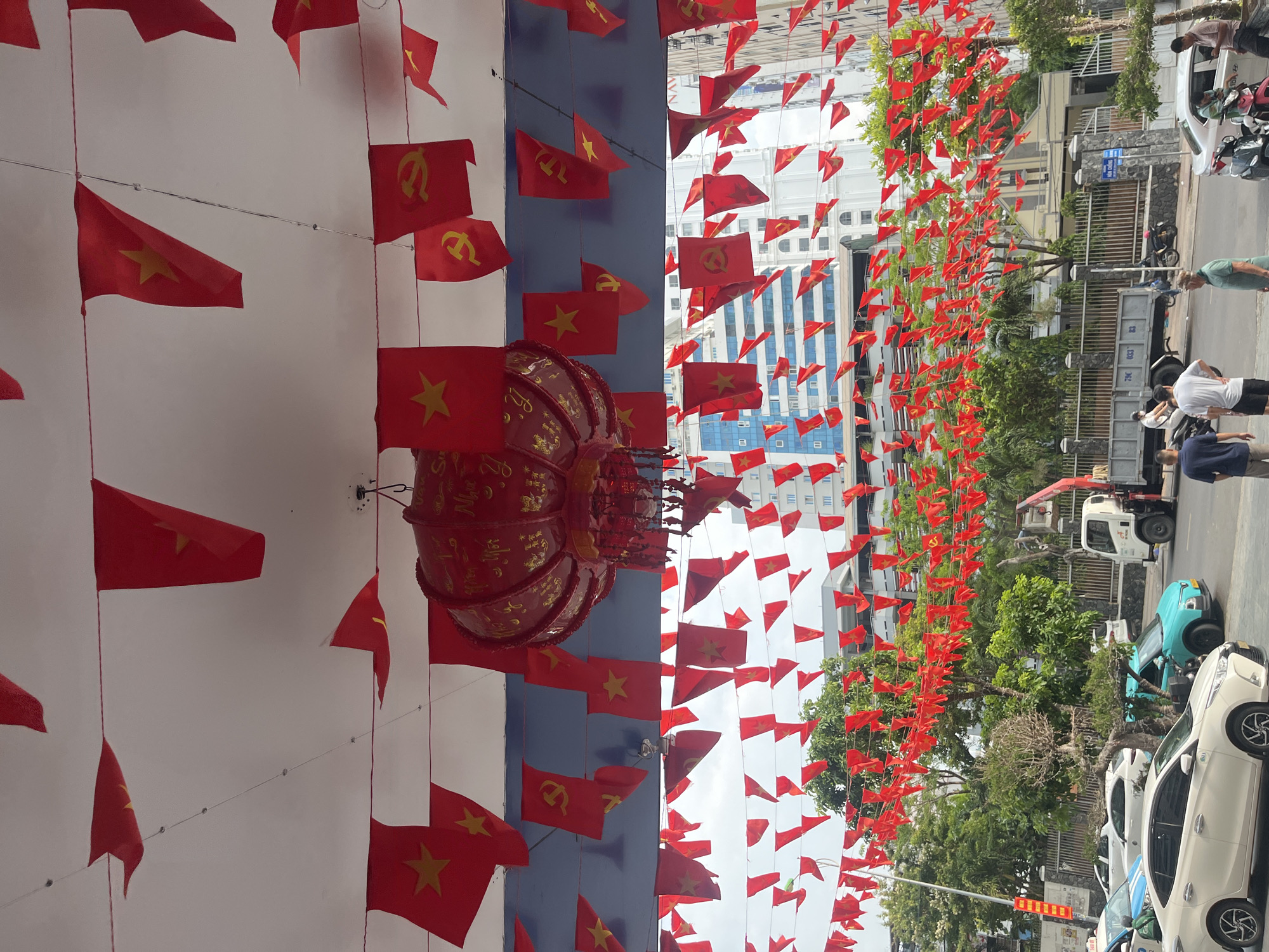 The image shows a street scene in Bac Nha Trang Ward, Vietnam, heavily decorated with Vietnamese flags. The flags are red with a yellow star. There is a red lantern hanging in the middle of the image. A street scene with cars, trees, and buildings is visible through the flags. People are visible walking on the sidewalk and standing near vehicles.