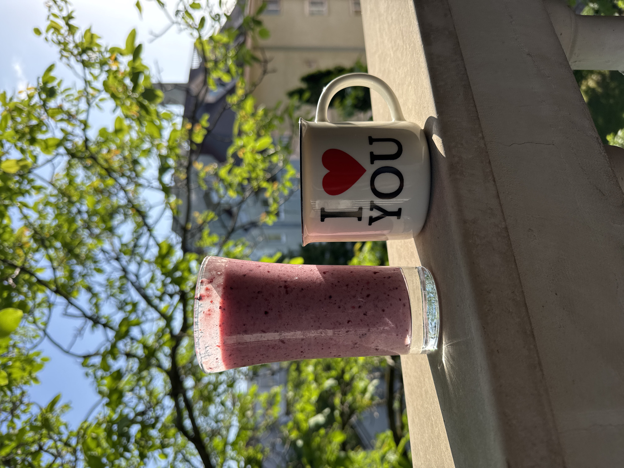 Here is a description of the image:
The image depicts a mug with the text "I (heart) YOU" and a glass of smoothie. These objects are placed on a white ledge, likely an outdoor balcony or railing. The background features blurred green foliage and a building in the distance, suggesting an urban or suburban setting. The lighting indicates it is daytime with bright sunlight casting shadows. The objects are static. There are no apparent people in the shot. The location is unspecified, as city and state are unknown.