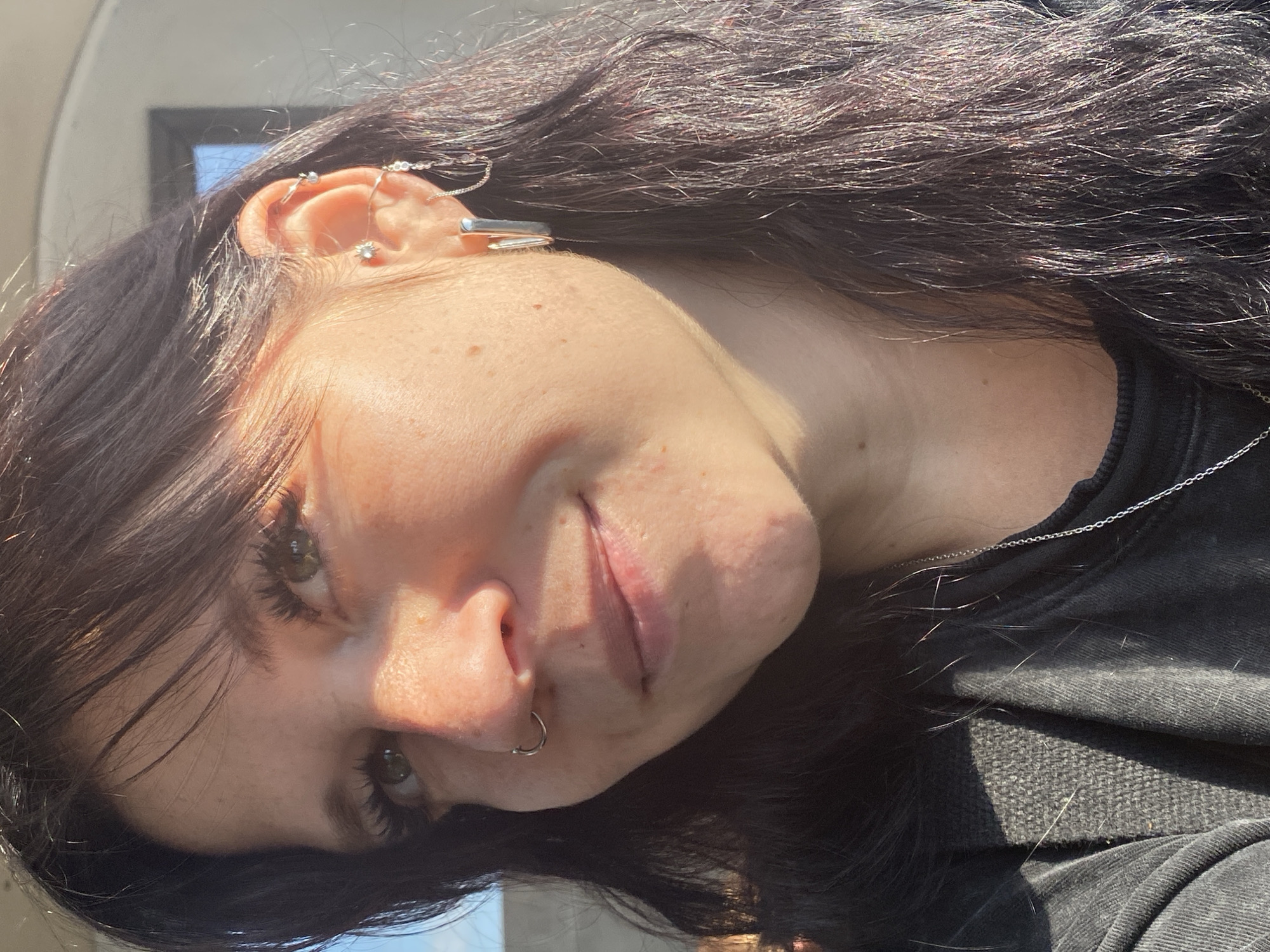 A close-up, waist-up, outdoor, natural light portrait of a young woman with dark hair and light green eyes. She is looking slightly to her right, with a subtle smile. The woman is wearing multiple earrings in her left ear, including a large hoop, a chain earring, and a stud. She also has a nose ring. Her hair is long and dark, with some strands catching the sunlight. She is wearing a dark grey shirt with a textured fabric on the shoulder area. The background is blurred, but appears to be an outdoor setting with hints of a building and sky. There are no specific landmarks visible to identify the location as Saint Petersburg, Russia.