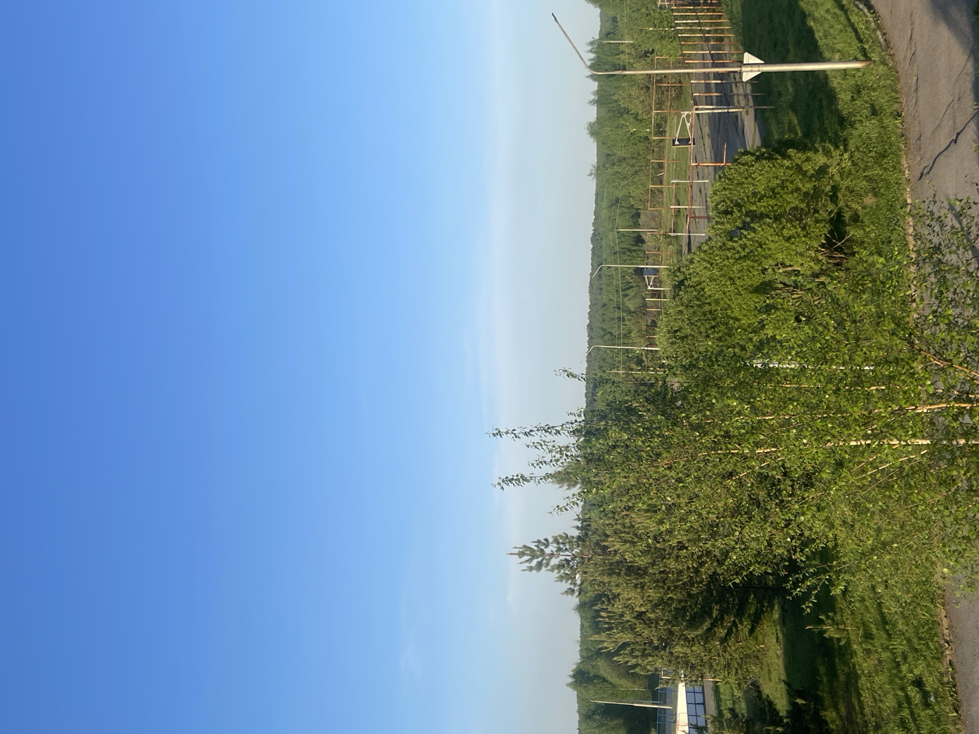 The image shows a landscape in Марковский, Russia, featuring a clear blue sky that dominates the left half of the frame. On the right side, there is lush greenery with various trees and bushes along a road. Utility poles and metal structures resembling fencing or barriers are visible. A segment of asphalt road is also present. The scene appears to be captured during daylight, likely a sunny day.