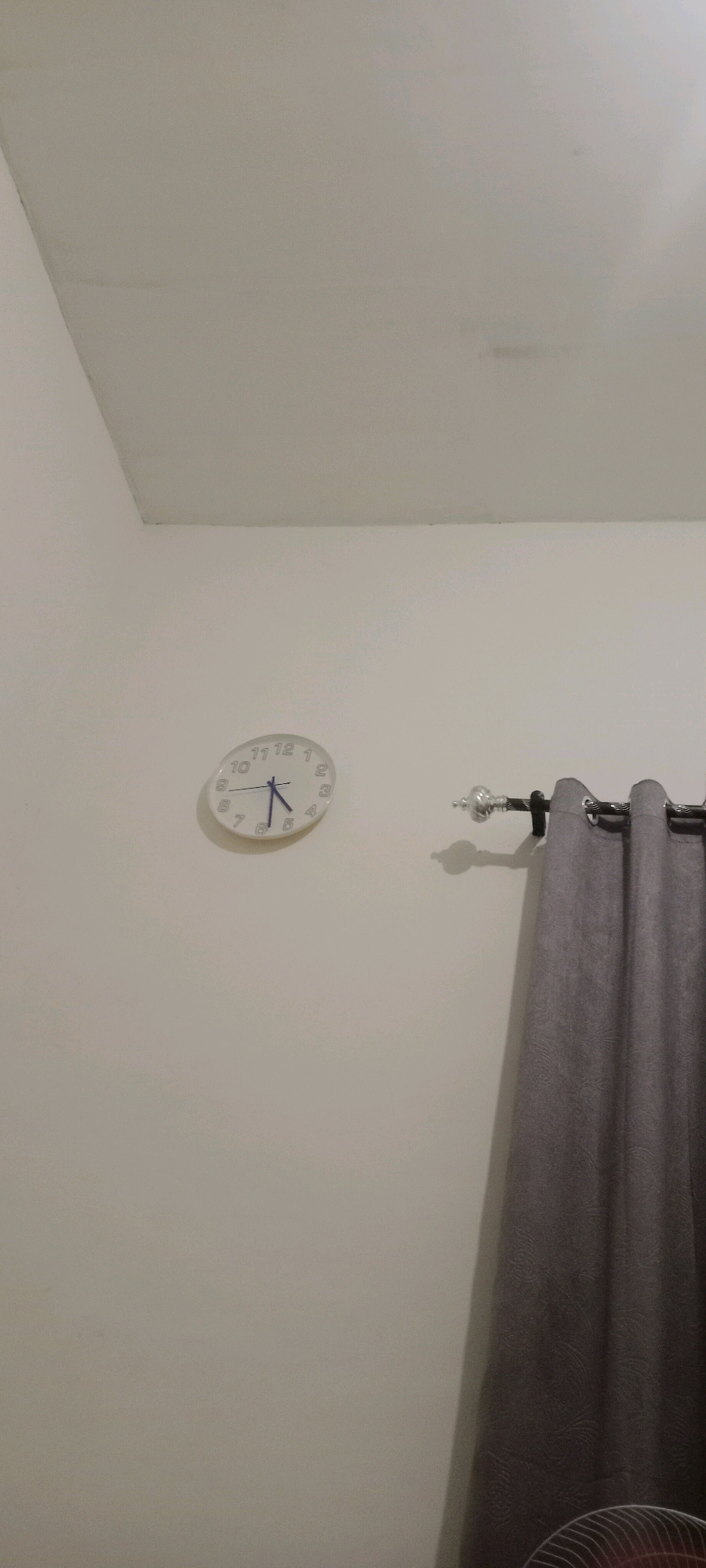 The image shows a white, oval-shaped analog clock on a light-colored wall. The clock face has black numerals and two hands: a blue hour hand pointing between the 5 and 6, and a shorter blue minute hand pointing at the 4. To the right of the clock, a dark grey curtain is partially visible, hanging from a curtain rod with decorative finials. The wall appears to be indoors, and there are subtle shadows cast on it. The ceiling is visible at the top of the image. Based on the provided information, the scene is located in Serongga, Indonesia.