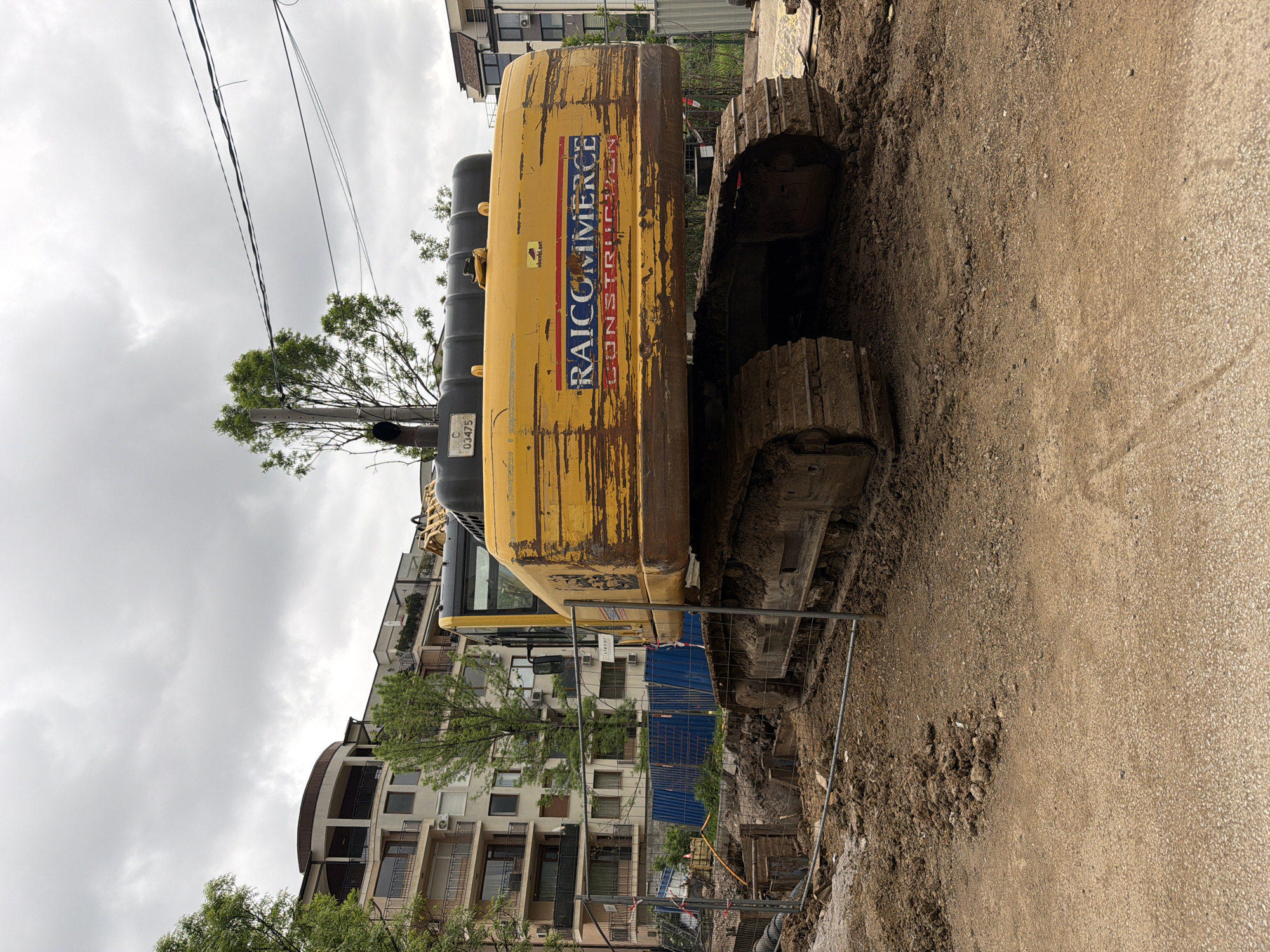 The image shows a yellow excavator with the branding "RAICOMMERCE CONSTRUCTION" parked on a dirt and asphalt surface, likely a construction site. The tracks of the excavator are muddy. A temporary metal fence separates the excavator from the construction area. In the background, there is a multi-story residential building, and utility poles with overhead wires are visible. The sky is overcast. A utility pole has "C 03475" written on it, suggesting that this is likely Bulgaria. Given the residential building style and the language on the excavator, it is very probable that the image was taken in Sofia, Bulgaria.