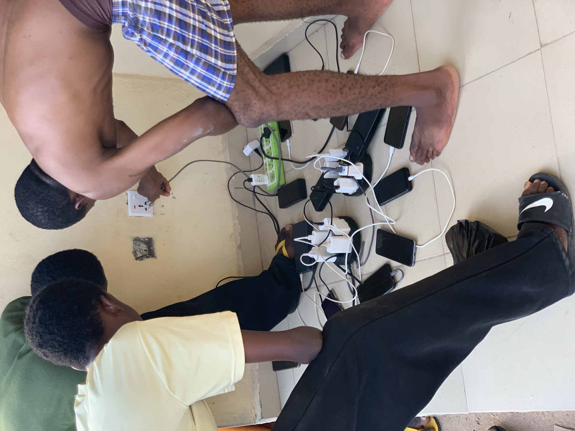 An overhead shot captures three individuals seated on a light-colored tiled floor, surrounded by numerous electronic devices undergoing charging. The scene is indoors, featuring plain walls.
In the upper part of the frame, a shirtless person wearing blue and white plaid shorts is actively plugging a white power adapter into a wall socket. The wall socket appears to be partially damaged or incompletely installed. This person's bare legs extend towards the central charging cluster.
The middle of the frame is dominated by a dense arrangement of charging equipment. Approximately eight smartphones and several power banks are scattered across the floor, connected by a tangle of white and black charging cables. These cables lead to multiple adapter bricks, which are plugged into two primary power strips: one green and one black multi-socket extension.
To the left, a person in a yellow t-shirt and black trousers sits with their hand positioned near the charging hub. Another individual, partially visible in a green top and dark trousers, is also seated nearby, observing the setup.
Footwear includes a black Nike-branded slide on the right side of the frame and a yellow slide partially visible near the charging devices. A black face mask is also on the floor, close to the charging hub. The individuals appear focused on the charging activity. The environment suggests an informal, communal charging area, potentially in Lokoja, Nigeria, as per the given context. No specific time of day or weather conditions are evident from the indoor setting. The only clearly visible text is the "Nike" logo on the slipper.