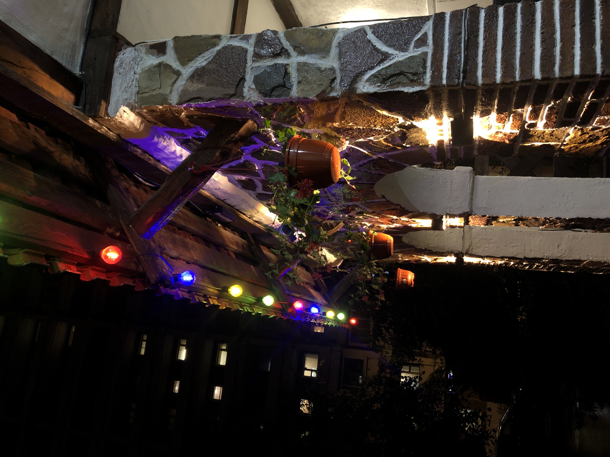 The image depicts an outdoor nighttime scene featuring an architectural structure adorned with colorful string lights and hanging potted plants. The structure is made of brick and stone, with wooden beams supporting a roof-like overhang. Several hanging plants in terra cotta pots are visible, some with red flowers. The string lights, in red, blue, green, and yellow, illuminate the scene, casting vibrant colors on the stonework and wooden beams. In the background, a dark building with illuminated windows suggests an urban setting. The overall impression is one of festive outdoor decoration.