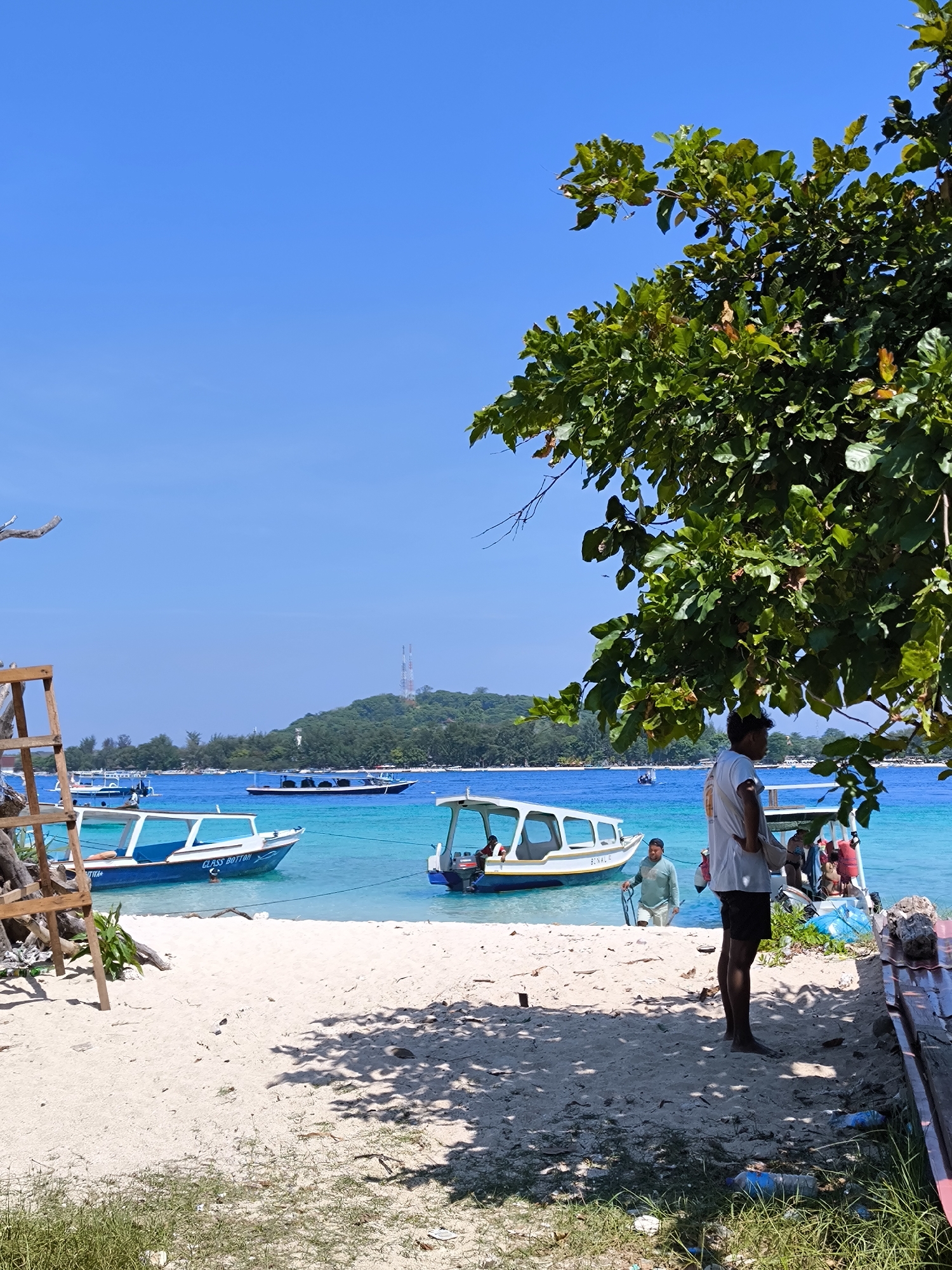 The image depicts a beach scene in Unknown, Indonesia, characterized by a vibrant turquoise sea under a clear blue sky. The foreground features a sandy beach with scattered debris, including plastic bottles and grass. To the left, a wooden structure, possibly a ladder or a support frame, is partially visible. Several boats are anchored in the shallow waters, varying in size and design; one is blue and white with the words "CLASS BOTTOM," and another is white with open sides. On the right, a man stands near a tree, his back to the camera, wearing a white shirt and dark shorts. Behind him, near the boats, other people are gathered, some wearing life vests. In the background, a green island with a communication tower on top is visible. The scene conveys a sense of relaxed tropical activity with evidence of human impact on the environment.