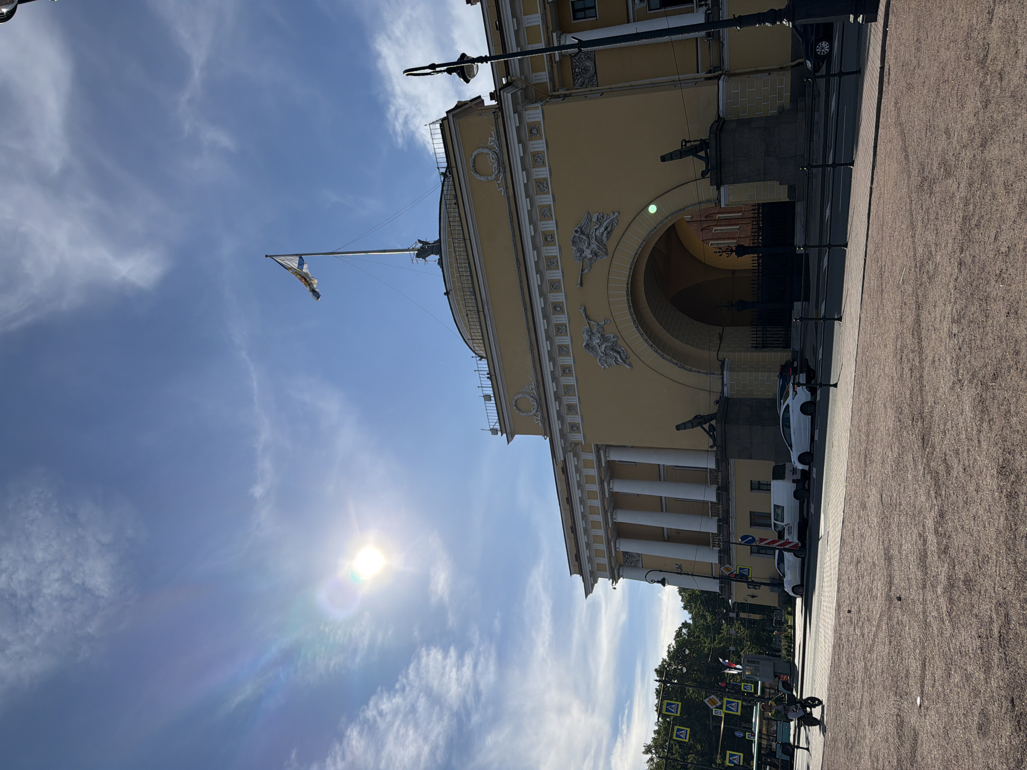 The image shows the Admiralty Building in Saint Petersburg, Russia, on a sunny day. The building, painted a pale yellow, features a large central archway, flanked by columns and adorned with sculptural reliefs of winged figures. A flag flies from a tall mast atop the building. In the foreground, a paved area leads to a street with several cars parked and driving. A lamppost stands to the right of the building, and trees are visible in the background to the left. A person on a scooter is seen in the mid-left of the frame, and pedestrian crossing signs are also visible. The sky is bright blue with scattered white clouds, and the sun is visible, causing a lens flare.