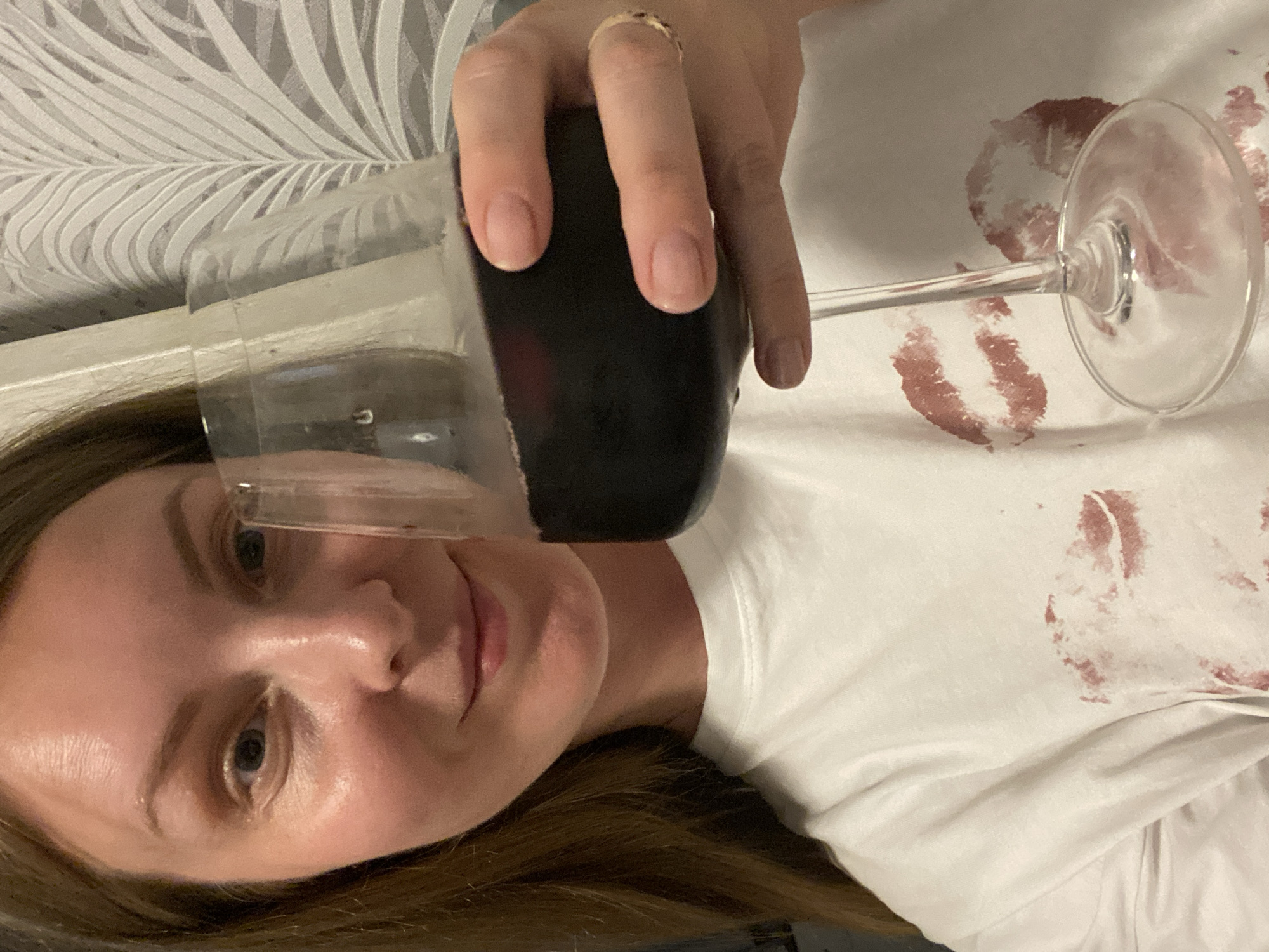 A woman is holding a glass of red wine. The glass is tilted and held close to her face. The wine appears to be dark red. She has light skin, light brown hair, and is wearing a ring on her finger. She is wearing a white shirt, and there are red lipstick stains on the shirt. The background includes a wall with a patterned wallpaper. The location is not specified in the provided image.