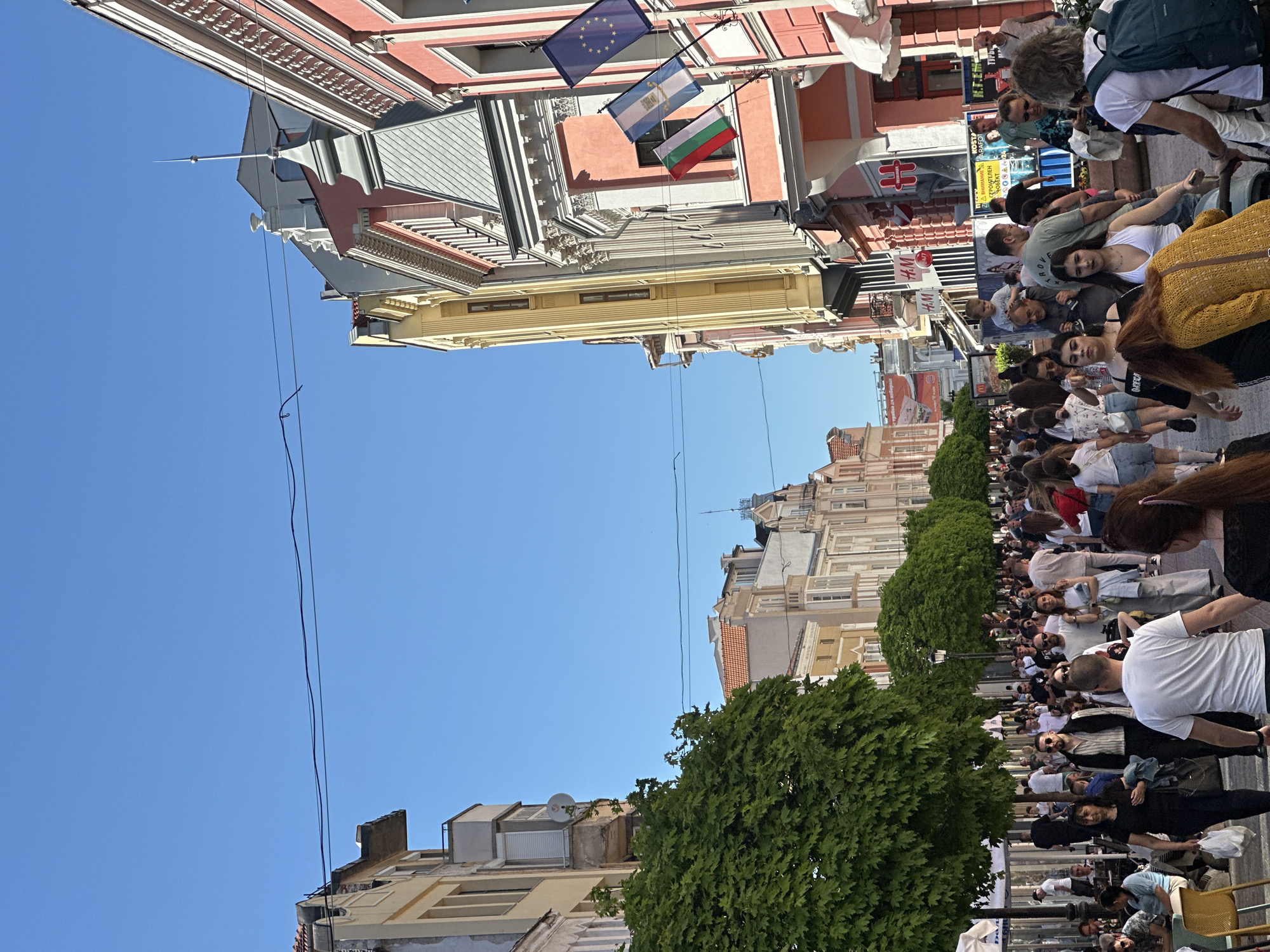 The image shows a street scene in Plovdiv, Bulgaria, with a large crowd of people. Buildings line both sides of the street, some displaying flags of the European Union, Bulgaria, and Argentina. The sky is clear and blue. Trees line the street, with electrical lines visible overhead. People of different ages and appearances populate the scene, many walking or standing. Several shops and stores are also visible.