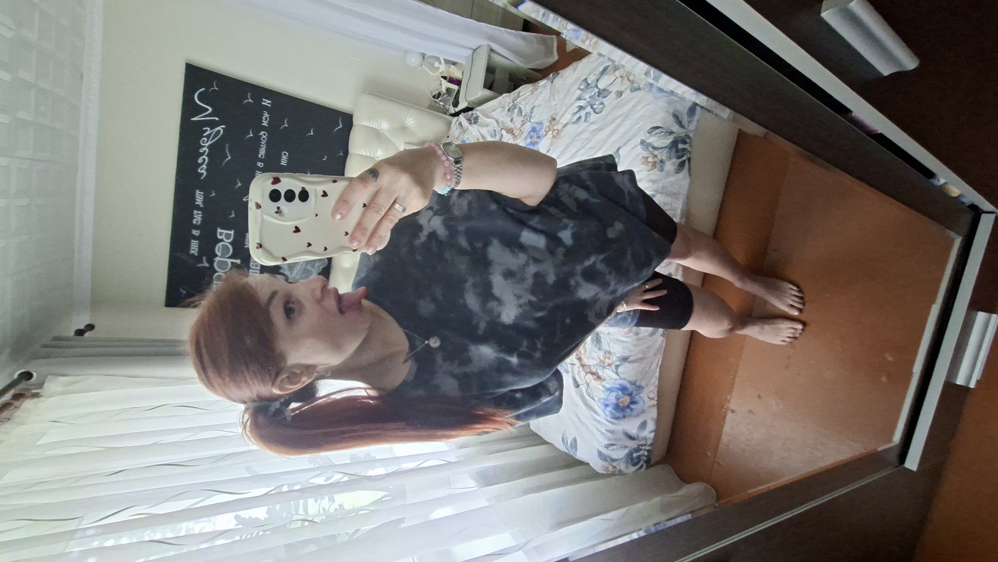 A woman with auburn hair in a ponytail takes a mirror selfie in a bedroom. She is wearing a dark, tie-dye t-shirt and black bike shorts. She has her tongue sticking out and is looking at her reflection. The room has a bed with a floral comforter, white curtains, and a mirror on the right side of the image. A framed picture with text is hanging on the wall behind the bed. A nightstand with a lamp and other items is visible next to the bed.