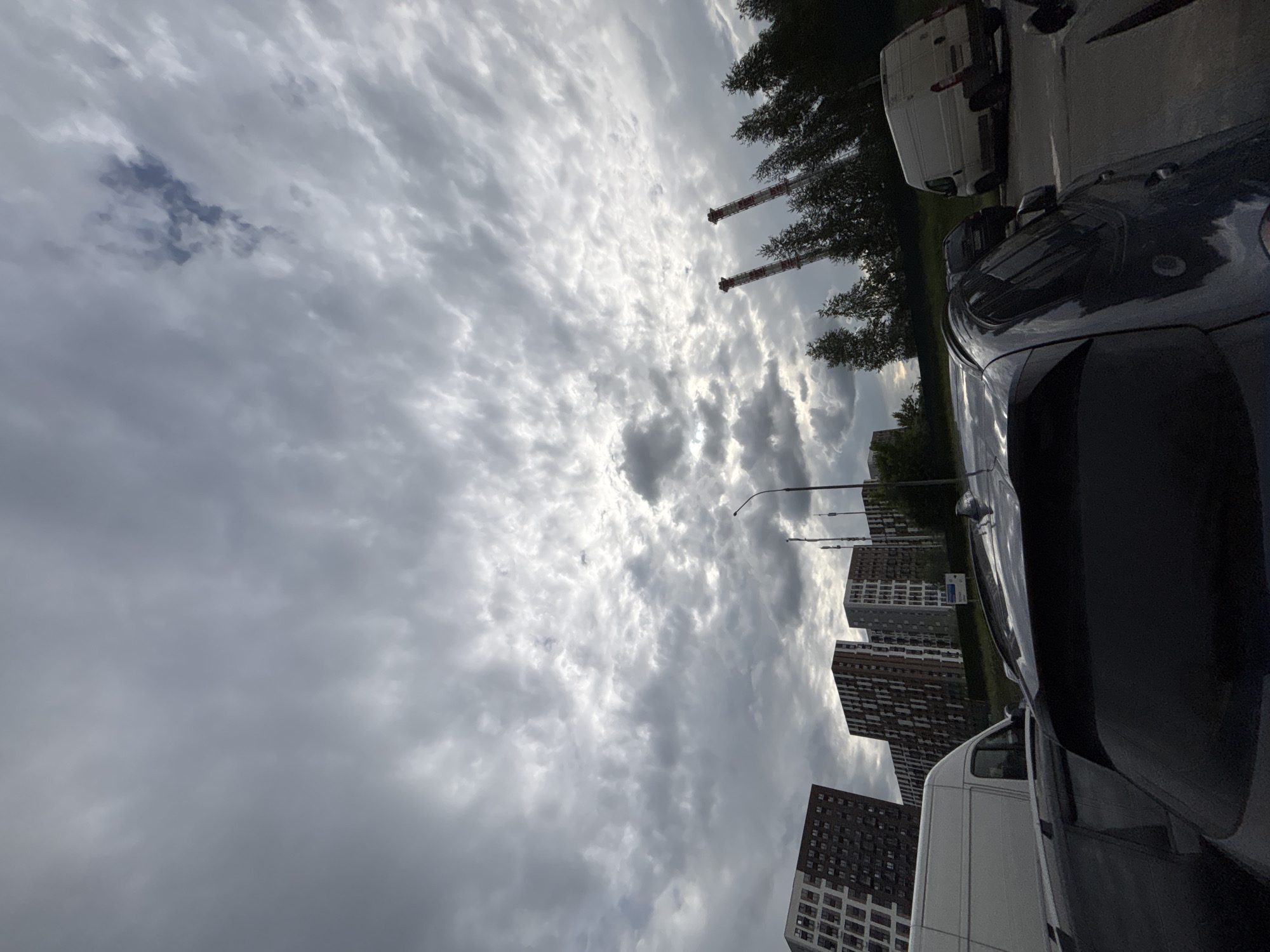 The image depicts an outdoor scene dominated by a cloudy sky. In the foreground, a dark-colored vehicle occupies a significant portion of the frame, with its reflective surface capturing the surrounding environment. Parked to the side of this vehicle is a white van. Further in the background, several tall residential buildings are visible, characterized by their multi-story structure and numerous windows.
The sky is filled with a variety of clouds, ranging from bright, illuminated patches to darker, more ominous formations. The lighting suggests it might be daytime, possibly late afternoon or early evening, given the diffused light. The presence of the buildings and parked vehicles indicates an urban or suburban setting. Two tall, industrial-looking structures with red and white banding are also visible in the distance, adding to the industrial or infrastructure element of the landscape. The overall mood is somewhat overcast and subdued. No people are visible in the image. No visible text is present.
