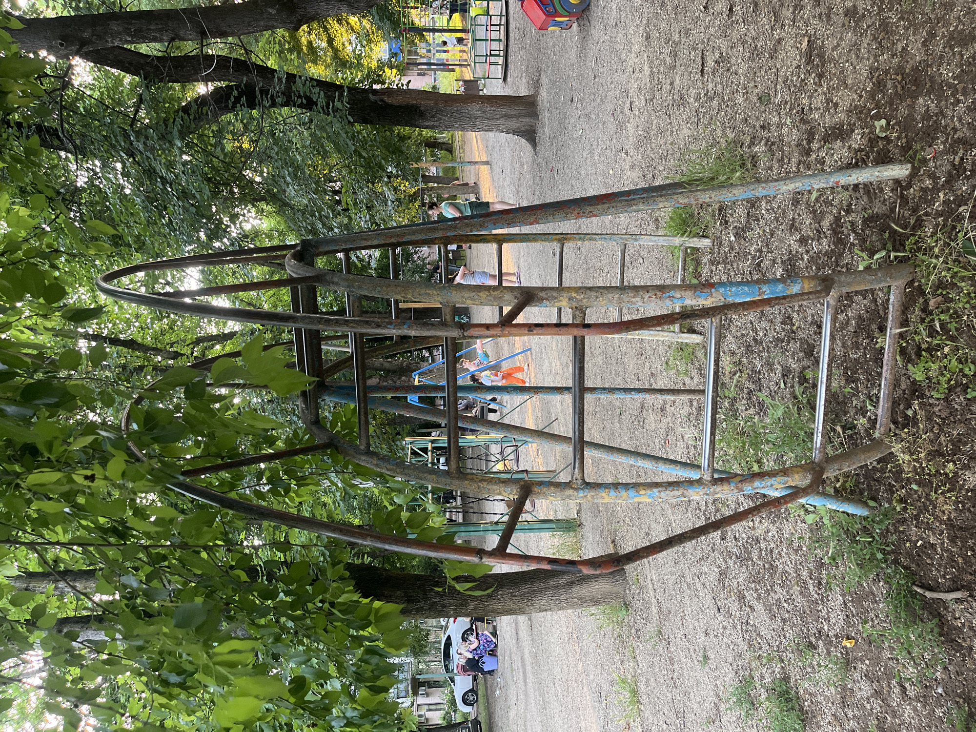 The image depicts a children's playground in Krasnodar, Russia, bathed in the soft light of late afternoon. In the foreground, a weathered metal jungle gym, painted in faded blues and reds, stands as the central subject. Its structure is a series of interconnected bars and ladders, showing signs of rust and wear. Sunlight filters through the lush green foliage of mature trees that encircle the playground, casting dappled shadows on the sandy ground.
In the background, the playground is sparsely populated. A woman in a green shirt and shorts stands with her arms crossed, observing the scene. To her left, another woman in a blue dress sits on a bench near a white car, accompanied by a child. Further into the playground, a few children are visible playing. One child in an orange outfit swings on a blue swing set, while another, wearing a white shirt and orange pants, walks near a merry-go-round. The environment is typical of an urban playground, with scattered gravel and patches of grass. The buildings in the distance suggest a residential area. The overall impression is one of a quiet, somewhat aged, but functional public space for children.