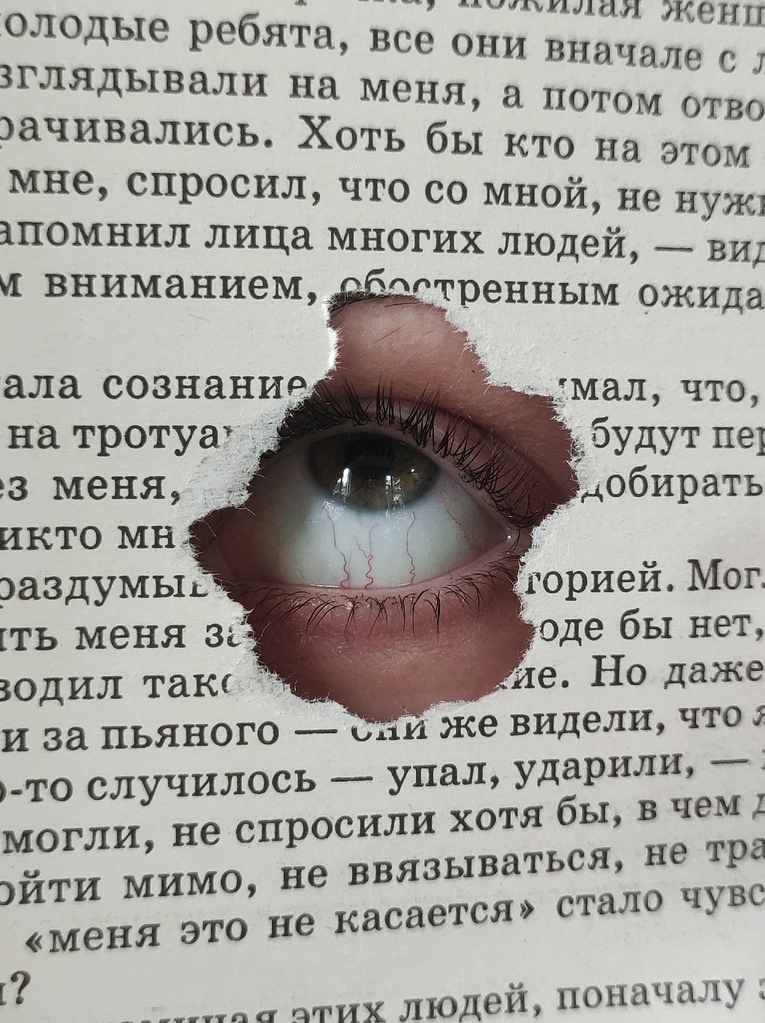 A close-up image shows an eye peering through a torn hole in a page of a book with Russian text. The visible text discusses observation, indifference, and personal responsibility. The eye is light-colored with visible blood vessels and eyelashes. The torn paper edges reveal the flesh tone of the surrounding skin. No location in the city of Фоки, Russia is provided or visible in the image.