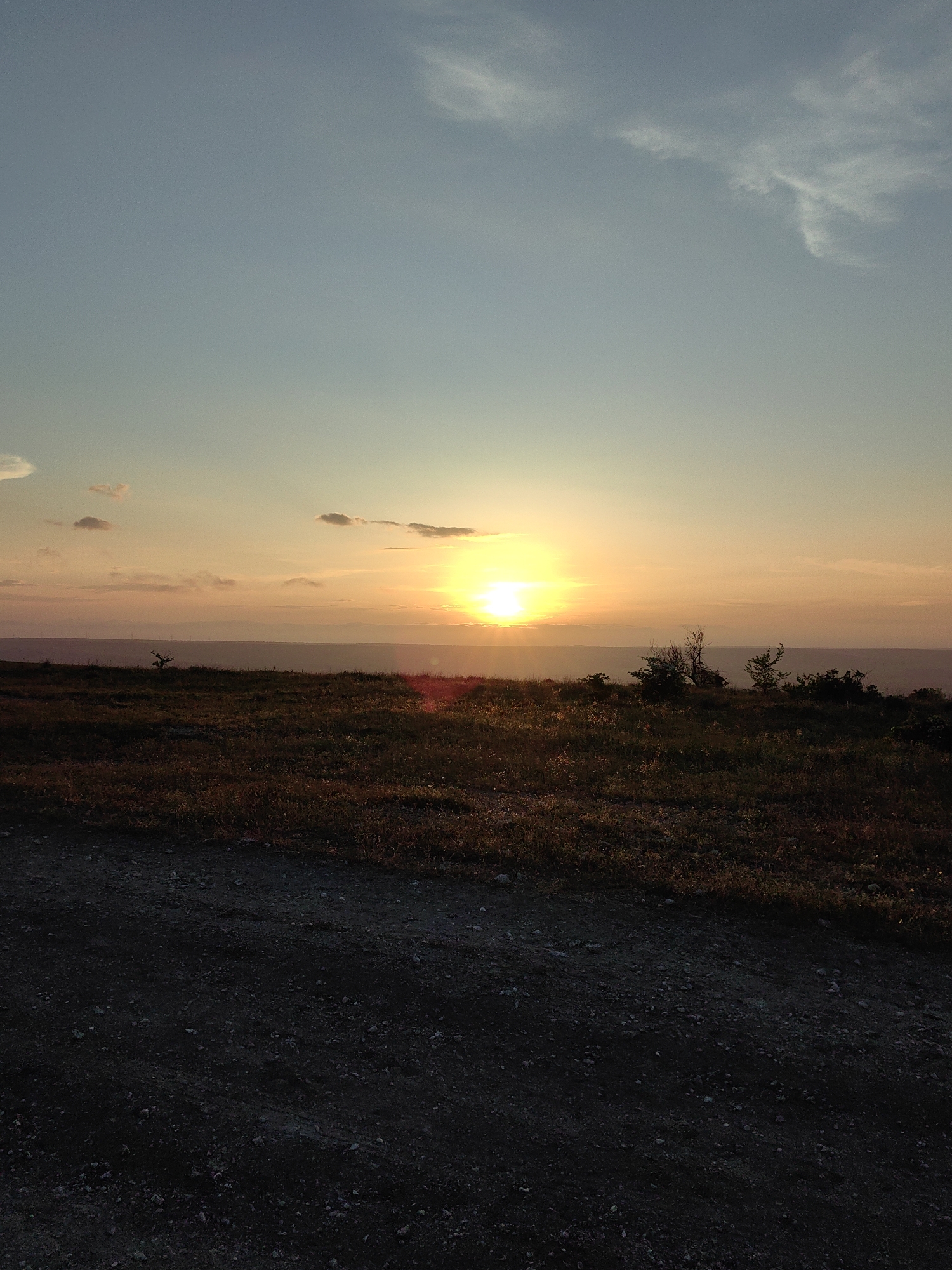 The image shows a landscape at sunset. The sky transitions from a light blue at the top to a warm orange near the horizon where the sun is setting. Thin clouds are scattered across the sky. Below the horizon, there is a dark, uneven terrain covered with low vegetation and small shrubs. In the foreground, there is a darker, gravelly area. The location is an open area in the city of Unknown, Unknown. There are no people visible in the image.