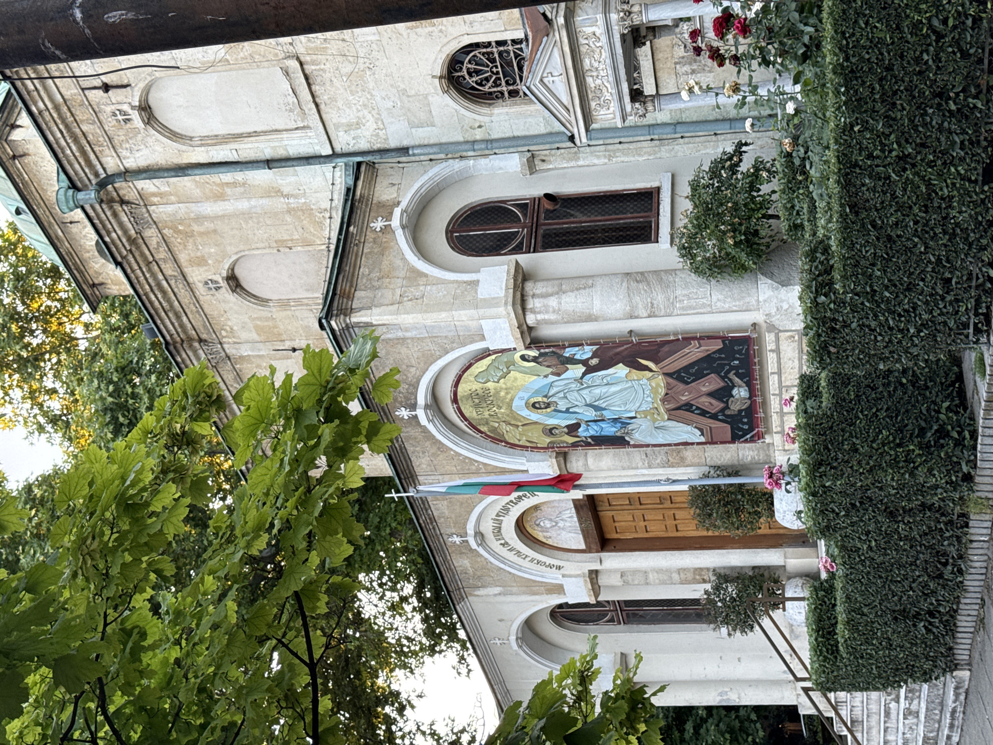 An exterior view of a stone building, possibly a church, in an outdoor setting. The building features arched windows, a dome, and a mosaic above the entrance. A Bulgarian flag flies on a flagpole to the left of the entrance. Lush green trees and bushes surround the structure, and rose bushes with pink and red blooms are visible in the foreground. A set of stone stairs leads up to the entrance. The lighting suggests it is daytime, possibly late afternoon or early morning due to the warm tones in the sky and foliage.