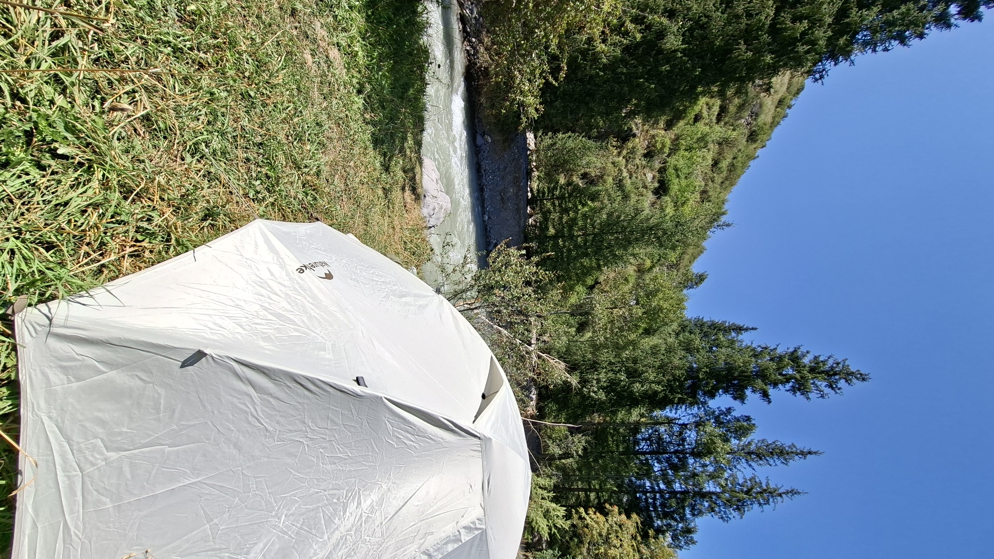 A white tent is pitched on a grassy bank next to a shallow, fast-flowing river in a mountainous, wooded area. The tent is in the foreground, taking up the right side of the image. The river is in the middle ground, flowing from left to right. Behind the river is a steep, wooded hillside with a clear blue sky above. The location is Batan, Kazakhstan.