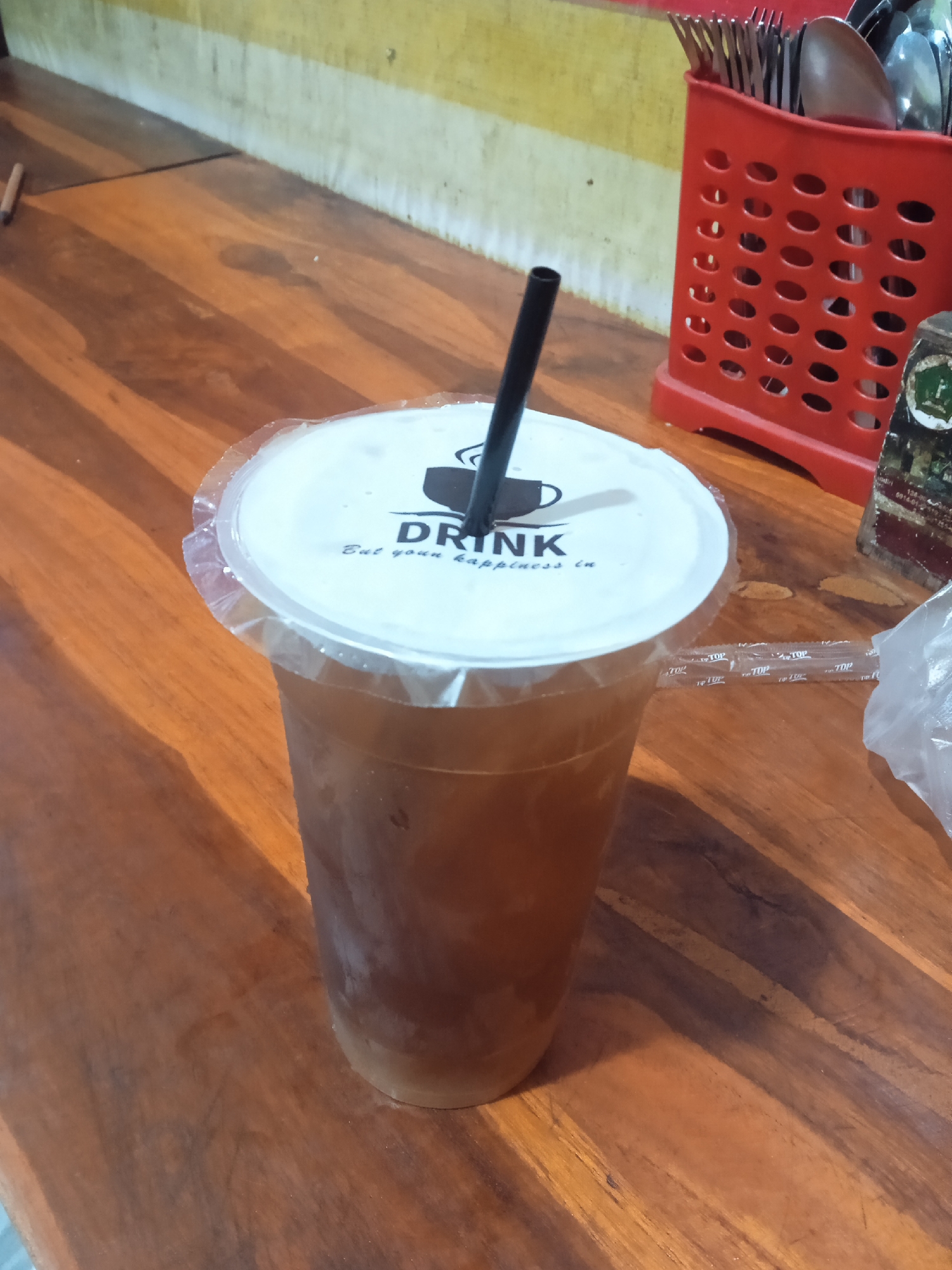 An iced beverage in a clear plastic cup with a white lid and black straw sits on a wooden table. The lid has the word "DRINK" printed in black and the phrase "Eat your happiness in" below it. The cup is filled with a dark liquid and ice. To the right of the cup is a red plastic utensil holder filled with spoons and forks, and a crumpled plastic bag. In the background, a yellow and white striped wall is visible. The scene is likely a cafe or restaurant in Semarang, Indonesia.