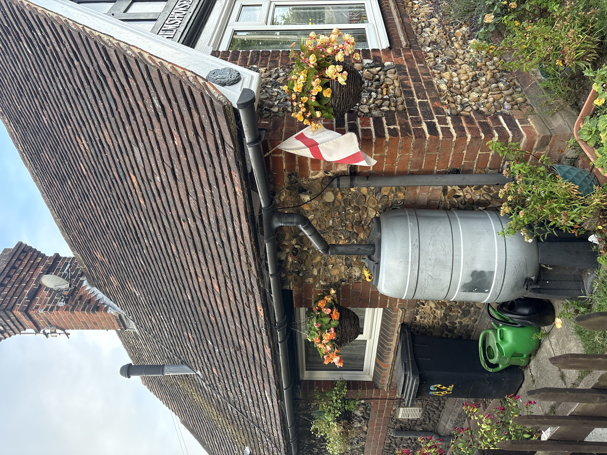 This image shows the side of a brick and flint building, likely a house, with a tiled roof. A downpipe from the gutter collects rainwater into a large grey water butt. Two hanging baskets filled with orange and yellow flowers are attached to the wall. An English flag hangs from the gutter. Next to the water butt is a black wheelie bin with the number "42" painted on it in colorful numbers, and two green watering cans and one black watering can. The building is located at Elmshouse, West Hanningfield Road, in the city of Chelmsford, United Kingdom. The garden area includes various plants and flowers. The sky is overcast.