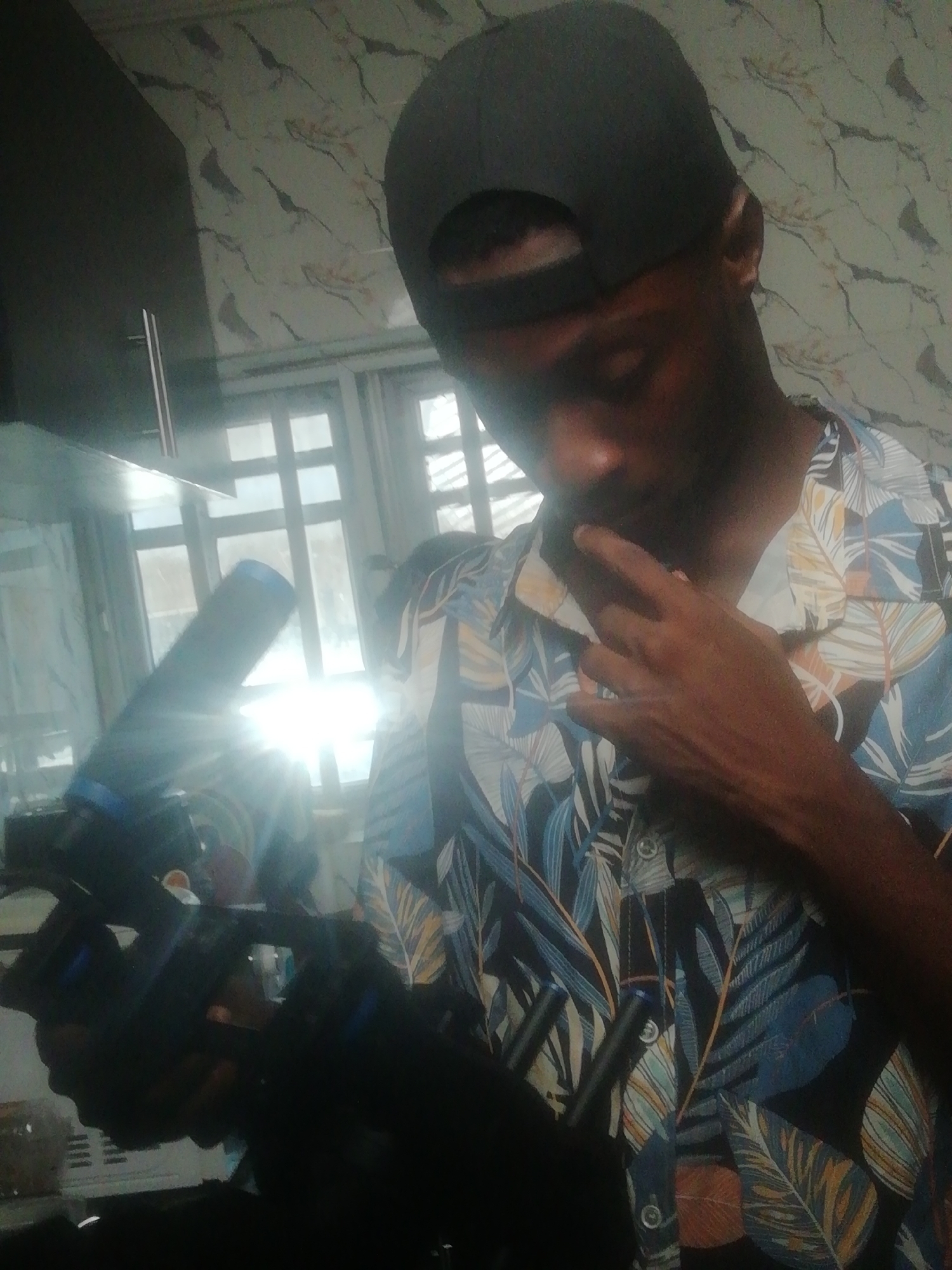 A person, likely male, is captured indoors in Ikorodu, Nigeria, under bright natural light. They wear a black baseball cap backward and a short-sleeved shirt featuring a tropical leaf pattern in shades of blue, black, white, and orange. The person's right hand is raised towards their chin or mouth in a gesture that suggests contemplation or focus.
In their left hand, the person holds a camera or video recording device, which is mostly dark but shows blue accents and is pointed towards a large, multi-paned window. Intense sunlight streams through this window, creating significant backlighting and strong shadows on the subject and the foreground, indicating it is daytime. The background reveals a textured or patterned wall/ceiling and a dark cabinet on the far left. No visible text is present in the image.