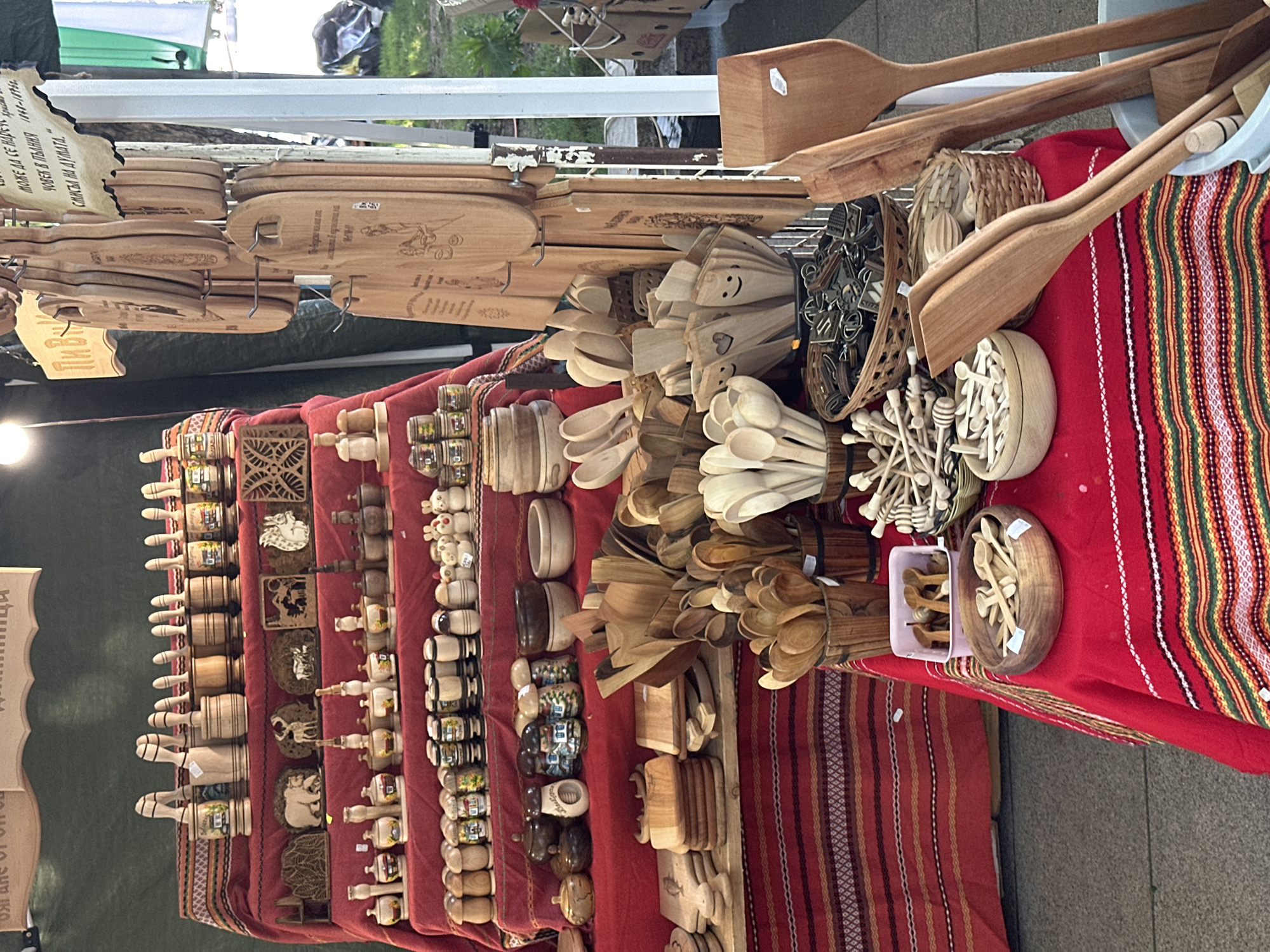 A market stall in Montana, Bulgaria, displays a variety of handmade wooden items on a red and striped tablecloth. The items include wooden spoons, spatulas, cutting boards, honey dippers, and small decorative figures. The stall is located outdoors, with a white metal shelf visible in the background.
