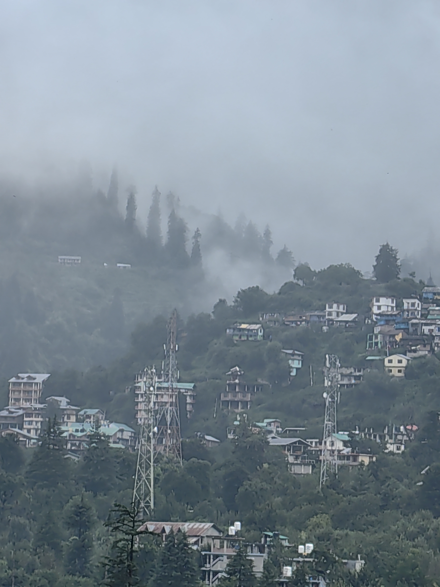 The image depicts a misty, overcast day in Manali, India, with the town built into a steep, green mountainside. The architecture is a mix of traditional hillside dwellings with colorful roofs and more modern multi-story buildings, including several with green-tiled roofs. A prominent cell tower stands near the center of the frame, surrounded by buildings and dense evergreen trees that cover the lower slopes. Further up the mountain, more structures are visible, some nestled amongst the trees and partially obscured by fog. The upper portion of the image is dominated by a thick layer of mist and fog that obscures the mountain peaks and creates a hazy, atmospheric effect. No people are visible in the image.