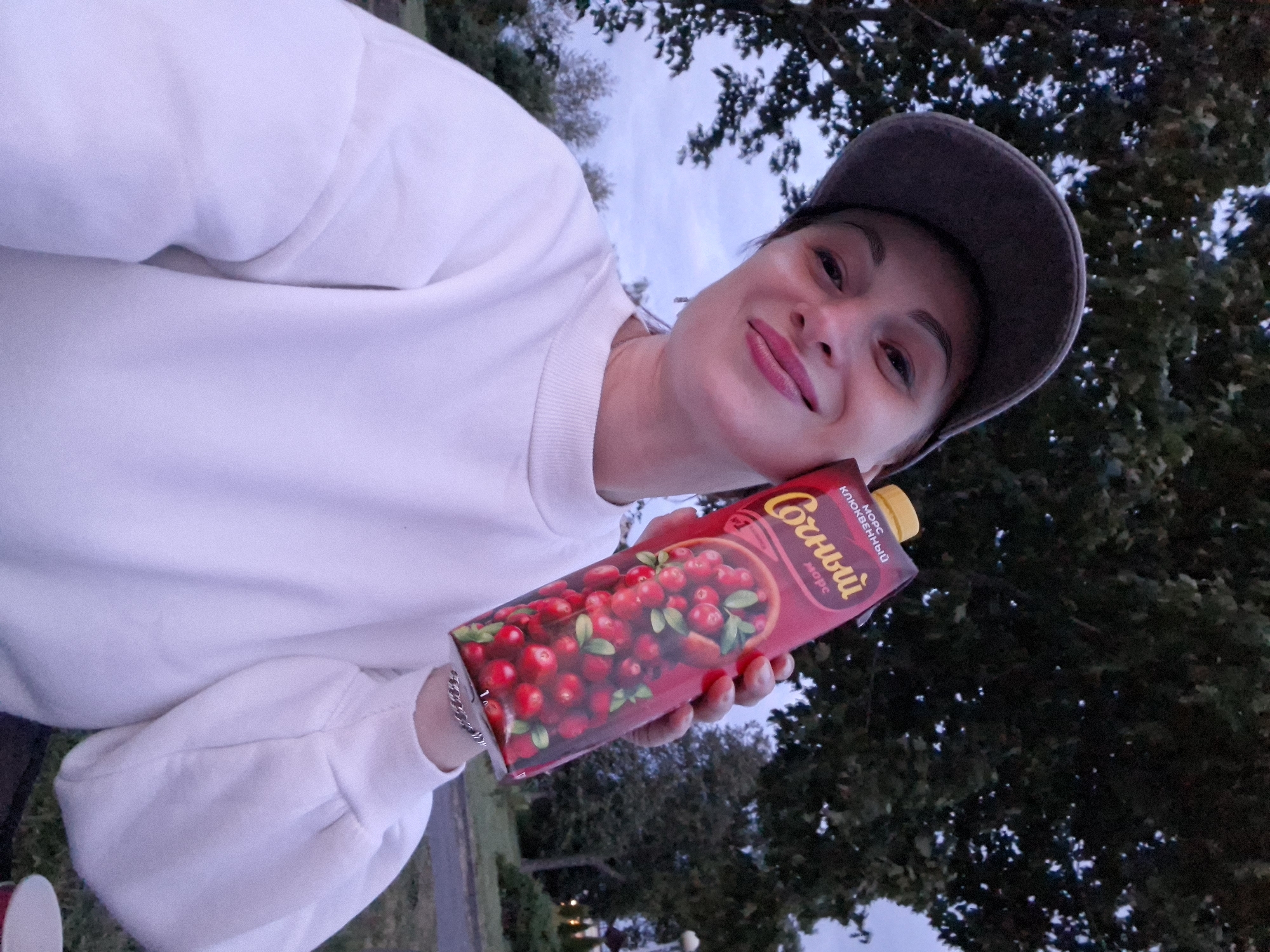 A woman wearing a white sweatshirt and a grey baseball cap holds a carton of cranberry juice in her right hand, partially obscuring her face as she takes a selfie outdoors. The carton displays a picture of cranberries and text in Cyrillic, including "Сочный" and "клюквенный". She is smiling and looking at the camera. The background suggests a park-like setting with trees and a cloudy sky, consistent with the environment in Myadzyel, Belarus. The lighting indicates it is dusk or early evening.
