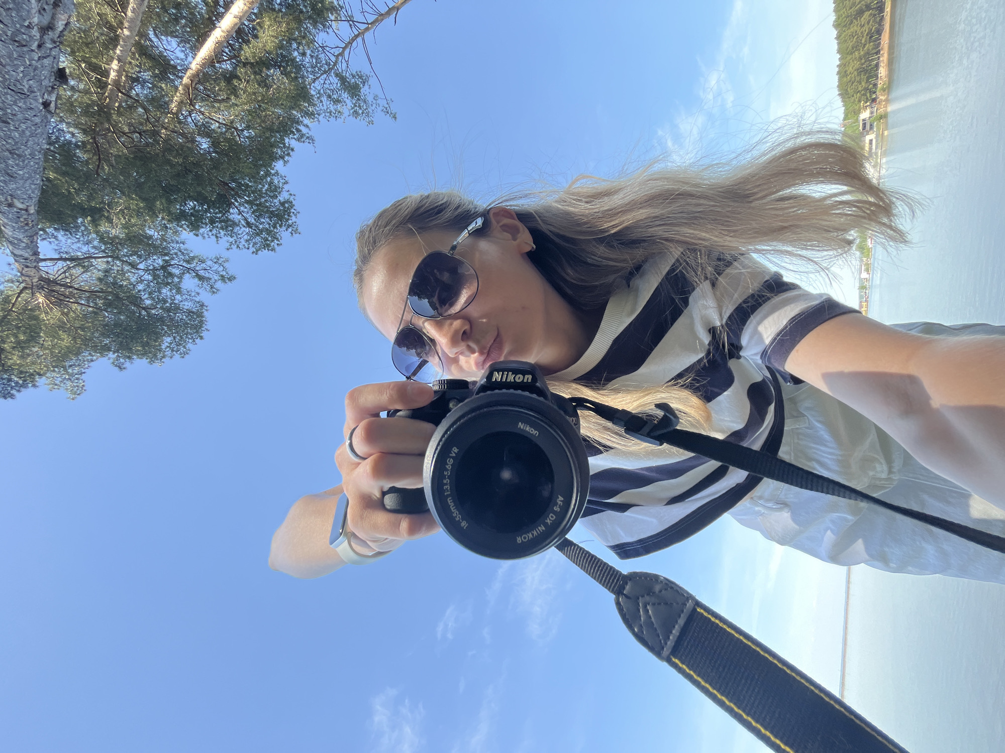 A woman wearing sunglasses and a striped shirt is holding a black Nikon camera, pointed towards the viewer. The background includes a blue sky, a body of water, and trees on the opposite shore. The scene appears to be near a body of water in Chaikovsky, Russia, based on the tree line, and other visual details such as building styles near the water. The camera lens is visible, and she seems to be taking a selfie. She is wearing a ring and a watch on her left hand. Her hair is light and blowing slightly in the wind.