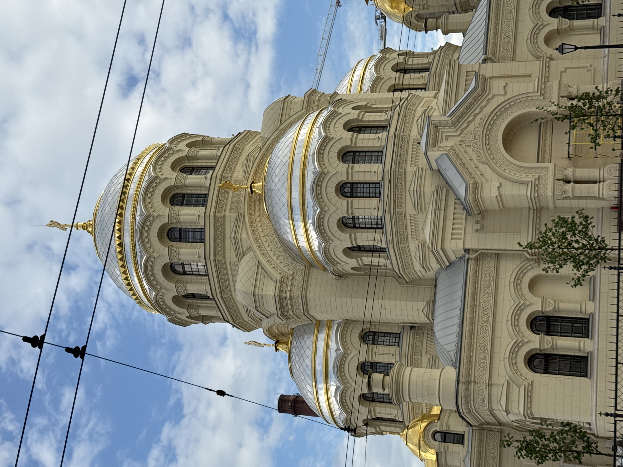 The image presents a grand architectural masterpiece, identified as the Naval Cathedral of St. Nicholas in Kronstadt, Saint Petersburg, Russia, under a bright, partially cloudy sky. The cathedral dominates the scene, showcasing its elaborate and ornate design.
The building is constructed from a light-colored material, possibly cream-colored stone or stucco, and is richly decorated. Multiple domes with a distinct Russian Orthodox style are visible; the most prominent dome in the foreground is large and gleaming silver, encircled by a golden band with intricate detailing, and topped with a golden cross. Several other smaller, golden-domed cupolas are scattered across the structure. The facade features numerous architectural details, including arched windows with dark panes, decorative columns, pilasters, and elaborate friezes with intricate patterns.
In the background, a construction crane can be seen, suggesting nearby development or ongoing maintenance. Overhead power lines or tram wires, with insulators, traverse the sky in the foreground. Towards the lower right, green foliage from trees or bushes partially screens a black metal fence, which appears to be adorned with cross-like finials. A lamppost and a yellow scaffold are also discernible near the building's base.
The scene is bathed in bright daylight, indicating it is likely late morning or early afternoon, with a clear blue sky interspersed with white clouds. There are no people visible, and no specific activity or event is taking place beyond the static display of the cathedral's grandeur. No visible text is present in the image.