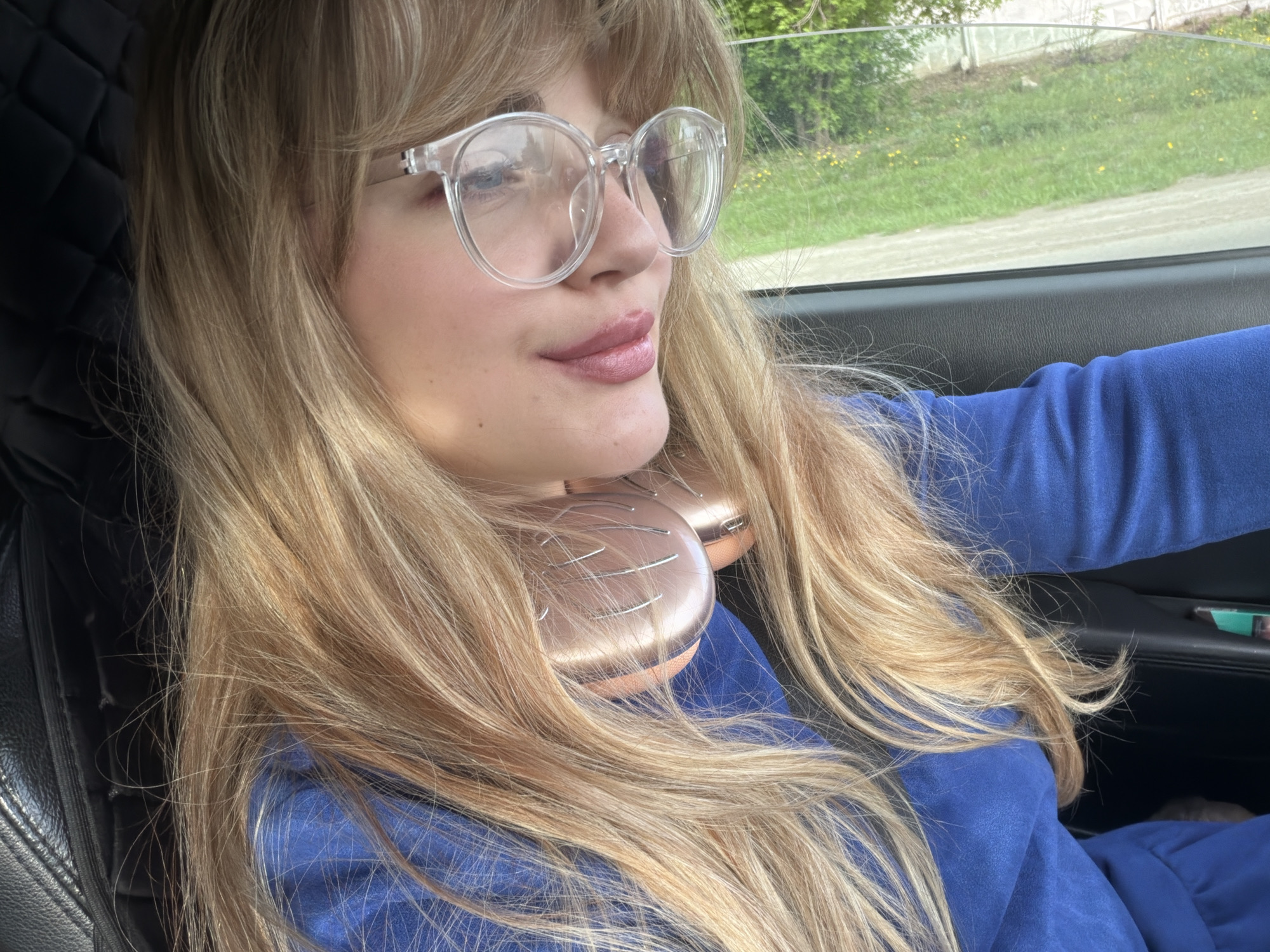 A woman with long blonde hair and clear-framed glasses is in a car. She is wearing a blue top. Headphones are around her neck. The car is traveling in Naberezhnye Chelny, Russia. The landscape seen through the car window appears to be a road bordered by grass and scattered vegetation. The car's interior is visible, including parts of the black dashboard and seat.