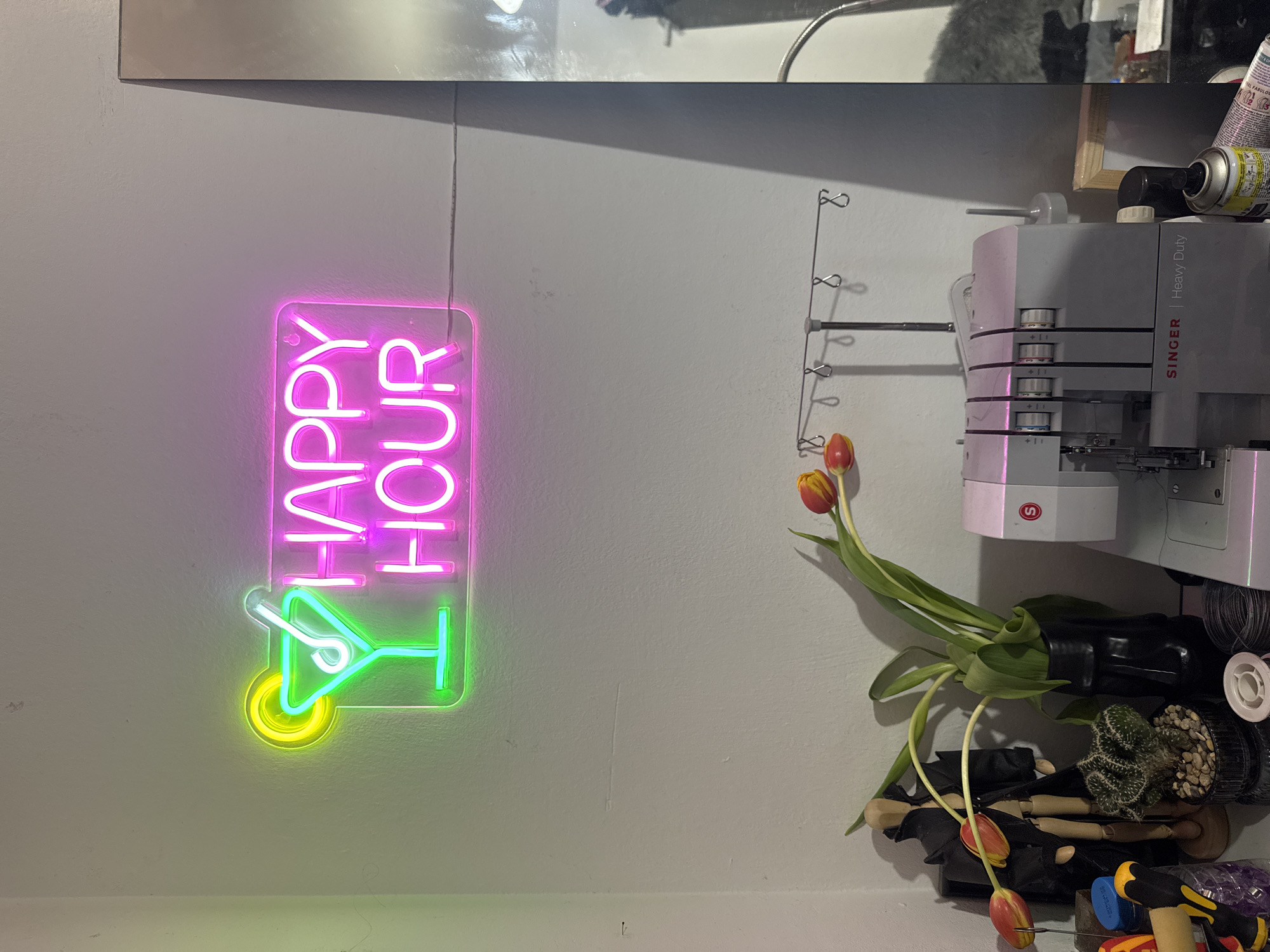 A vertical, medium shot depicts a segment of a wall in an interior setting. A "HAPPY HOUR" neon sign, illuminated in pink and green, is affixed to the wall, the sign's cocktail glass design in green with a yellow base. To the right of the sign, a metal rack with several looped hooks extends downwards. A small bouquet of tulips, predominantly orange with green stems, is arranged next to the rack. Further to the right, a Singer heavy-duty sewing machine is visible. Below the tulips and the sewing machine, a black vessel holding a cactus and other items is situated. Additional objects, including tools and containers, are present at the base of the scene. The wall is a neutral color, likely white or light grey. The scene appears to be indoors, likely a workshop or craft room, as suggested by the sewing machine and tools. There is no indication of Sofia, Bulgaria.