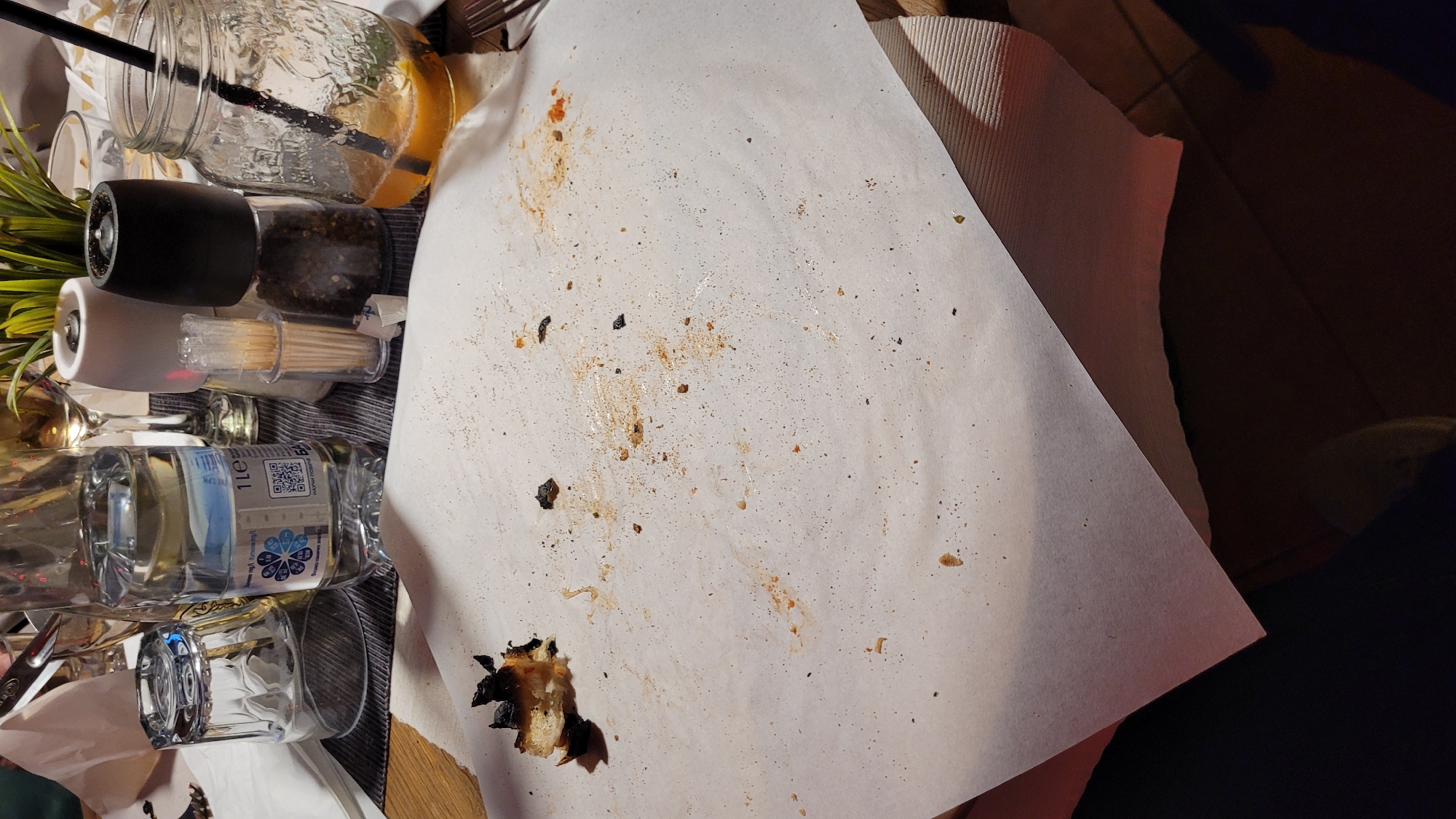 The image captures a detailed overhead view of a table setting, likely after a meal. The scene is dominated by a crumpled, grease-stained white paper placemat in the center and right, covered with various food residues including orange and brown sauce splatters, small dark specks, and larger burnt crumbs, with a significant piece of charred bread or crust in the bottom left.
To the left of the paper placemat, several dining items are arranged. These include a clear glass jar with a black straw and a lemon slice at the top, a black pepper grinder, and a white salt grinder directly below it. Next to the grinders is a clear container filled with numerous wooden toothpicks. Further down on the left, a transparent plastic bottle of water is visible, clearly labeled "1Le" (1 liter) and featuring a QR code. Below the QR code, the text "Марица" (Maritsa) is discernable, indicating a Bulgarian brand, which aligns with the provided location context of Varna, Bulgaria. A blue and white stylized flower or star logo is also present on the bottle. An empty, ribbed glass tumbler stands beside the water bottle. A green artificial plant provides a touch of decor on the far left.
An underlying striped beige or light brown placemat or napkin can be seen beneath the white paper and other items. The background to the far right is dark and indistinct, possibly a shadowed floor or chair. The lighting is artificial, suggesting an indoor setting, potentially in the evening. No people are visible, and no specific activities are taking place, but the remnants on the placemat strongly indicate a recently concluded dining experience.