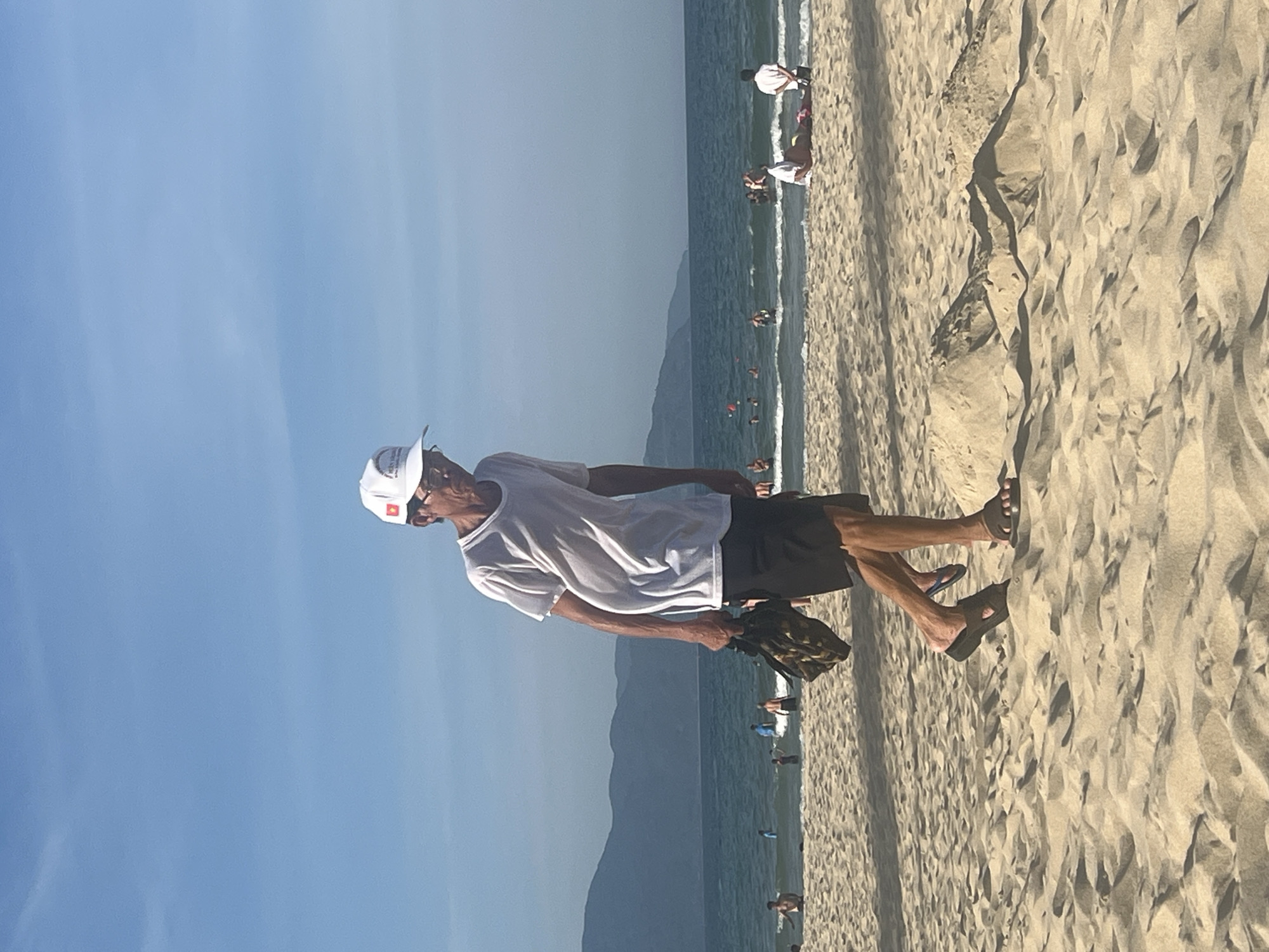 This is a medium shot photograph taken on a sandy beach in Đà Nẵng, Vietnam. The foreground is dominated by a man walking left to right across the frame. He is wearing a white t-shirt, black shorts, a white baseball cap with a red emblem, and dark sandals. He is holding a bag with a camouflage pattern in his right hand. His face is visible, and he is looking down as he walks.
In the background, the ocean is visible with gentle waves. Several people are in the water and on the beach. A distant mountain range forms the horizon. The sky is a clear blue with some faint clouds. The overall impression is a sunny day at the beach with recreational activities taking place.