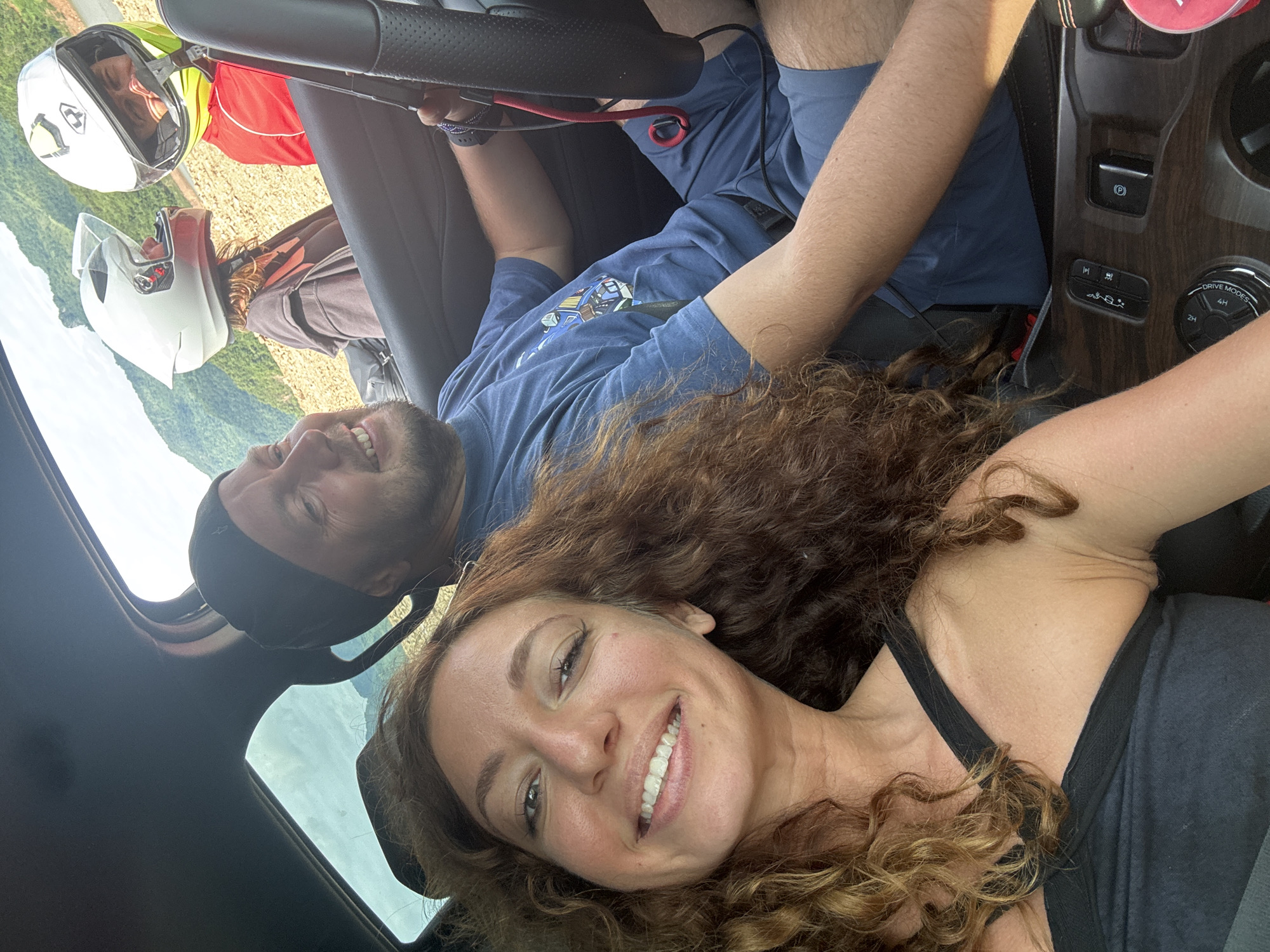 In the city of Tân Phong, Vietnam, a selfie is taken inside a vehicle with a landscape background of lush green mountains and a body of water. The driver, a man with a beard and a black beanie, smiles as he holds the steering wheel. Beside him, a woman with curly brown hair and a black tank top beams at the camera. In the back seats, two individuals are visible, both wearing motorcycle helmets - one in white and the other in white with a yellow visor. The interior of the car shows a dashboard with various controls and a center console. The overall scene suggests an adventurous outing or travel.