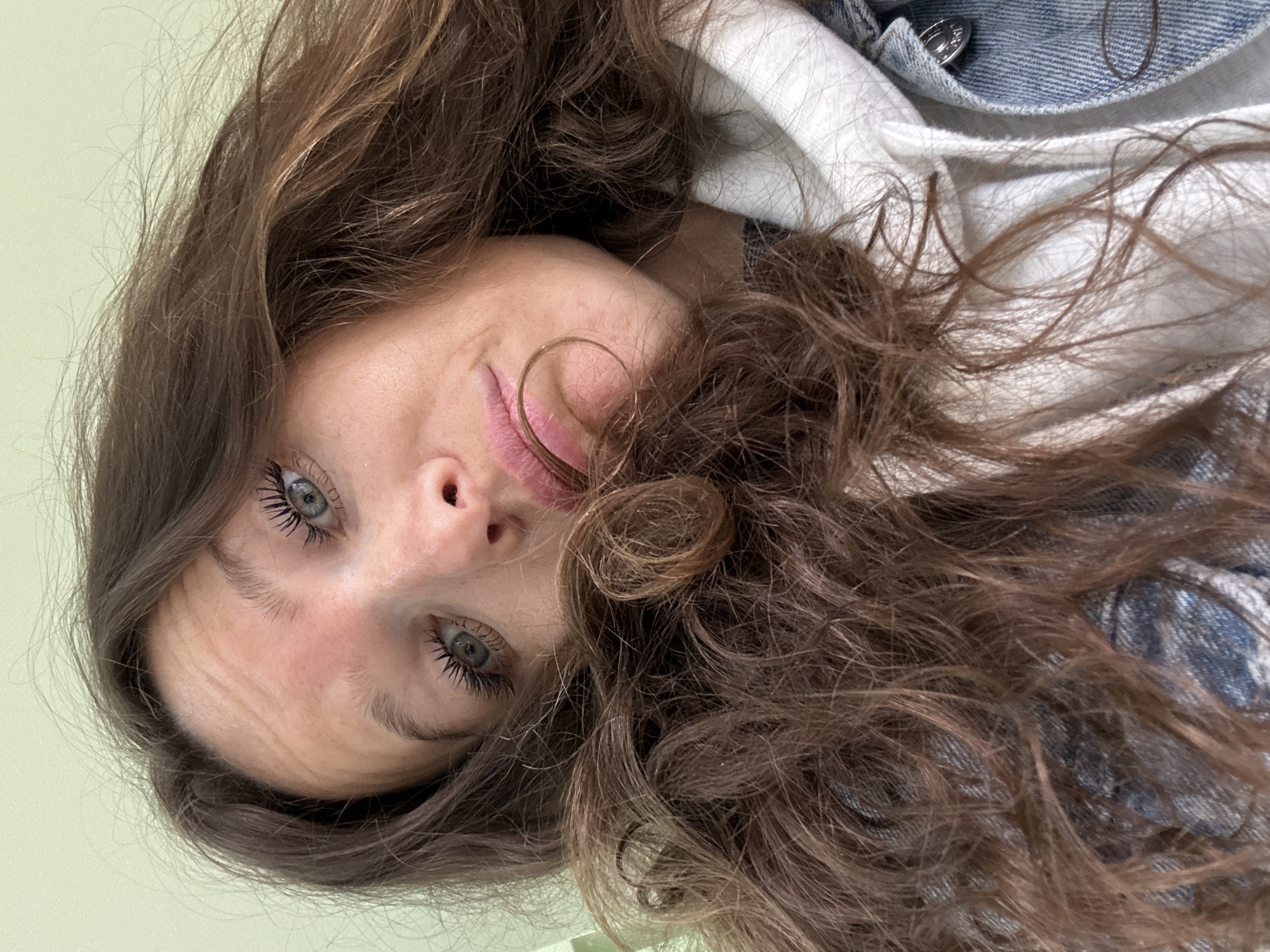 The image is a close-up, low-angle selfie of a woman with long, wavy brown hair and light-colored eyes. She is wearing a denim jacket over a white hooded sweatshirt. Her hair obscures a portion of her face, with strands framing her eyes and mouth. The background is a plain, pale green wall. No specific location in Moscow, Russia, can be determined from the image.