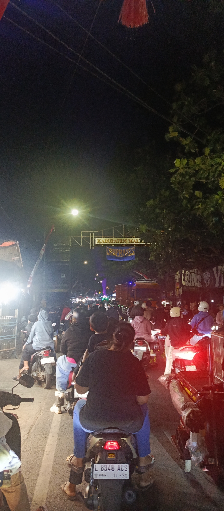 The image captures a nighttime scene on a street in Sekarpuro, Indonesia, likely during a traffic event. A dense crowd of people on motorcycles is depicted, creating a traffic jam. The foreground shows the backs of several individuals riding motorcycles, with one license plate clearly visible reading "L 6348 ACS". The motorcycles are of various models and colors, with some having visible rear brake lights illuminated. In the background, a gate or archway is visible with text that appears to say "KABUPATEN MALI". There are also trees lining the street, and streetlights contributing to the ambient illumination. The overall atmosphere suggests a busy and crowded urban environment at night, possibly a celebration or event that has led to a significant gathering of vehicles.